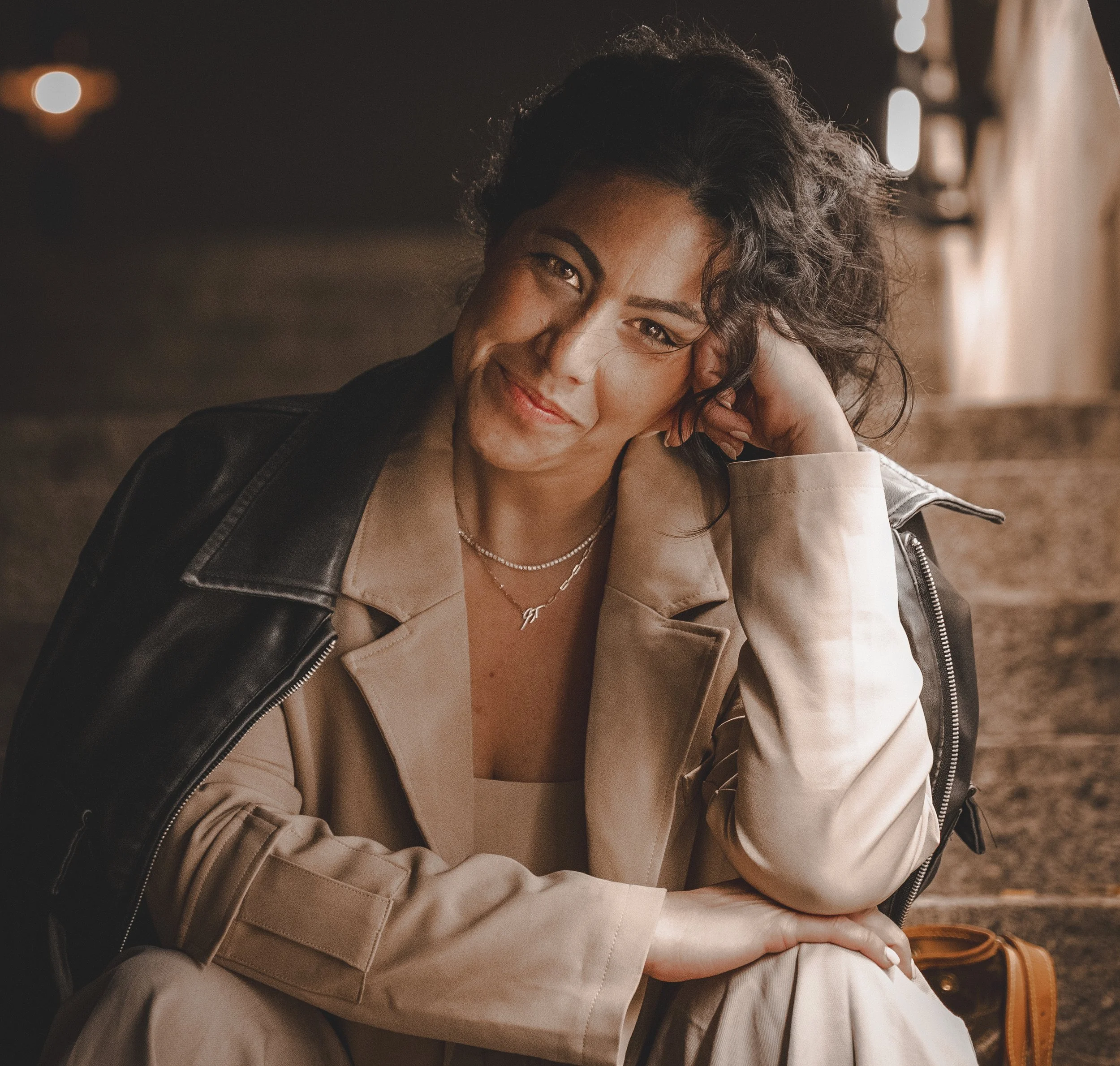 A woman with dark curly hair, wearing a beige coat over a light-colored outfit, sitting on stairs with her head resting on her hand, smiling at the camera. She has layered necklaces and a black jacket draped over her shoulder.