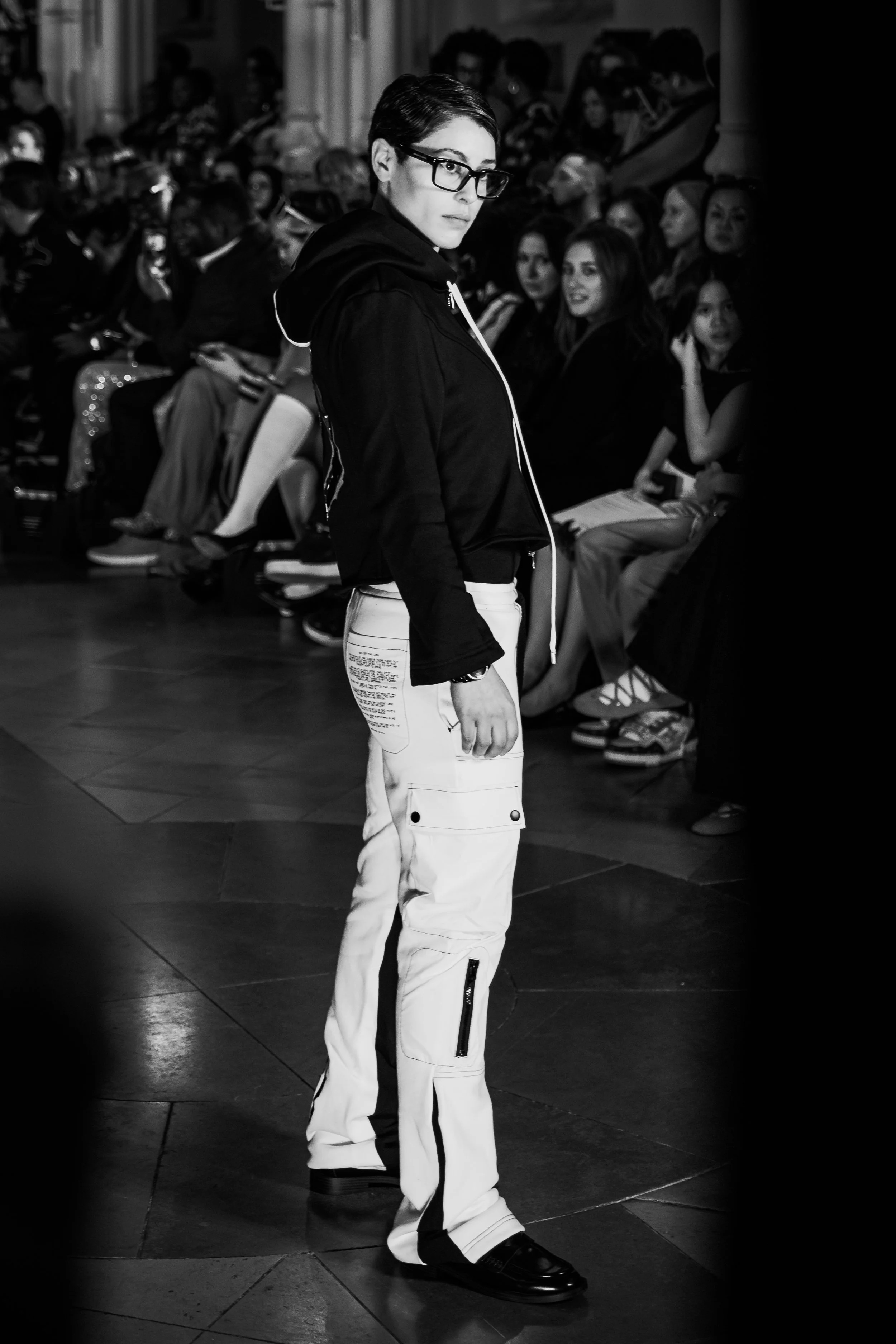 A young woman with short hair and glasses standing in an indoor setting, possibly a runway or fashion show, with an audience sitting in the background.