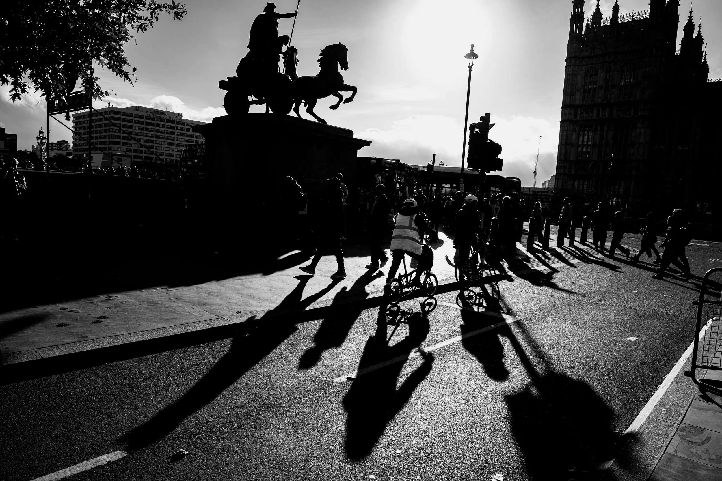 Silhouettes of pedestrians walking and cyclists riding past a monument of a chariot with a horse and a figure holding a spear, with a Gothic-style building and a bus in the background, in black and white.