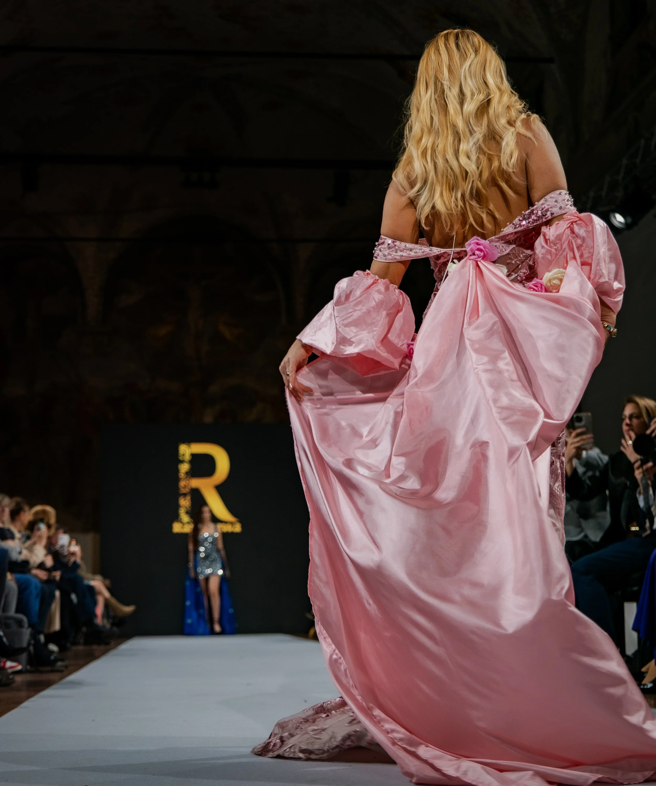 a female model walking down a runway wearing a pink gown with large bow details, onlookers and photographers on either side, and a black background with a large yellow 'R' behind her.