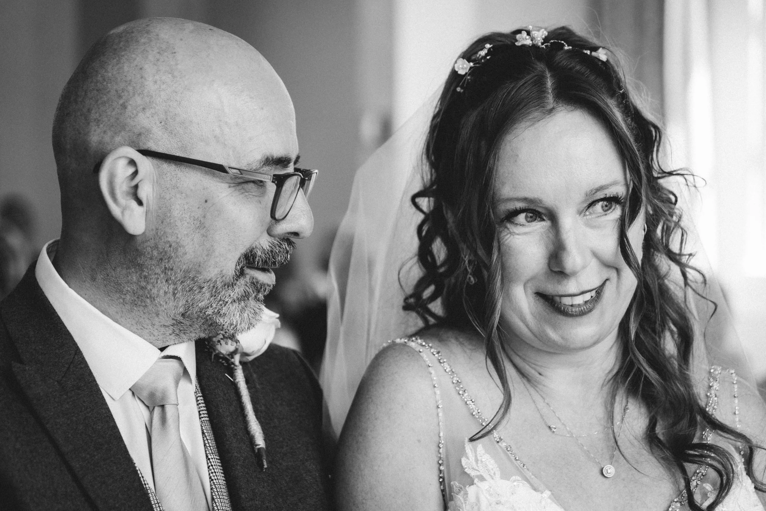 A black and white photograph of a bride and groom, with the bride smiling and looking slightly to the side and the groom looking down, both dressed in wedding attire.