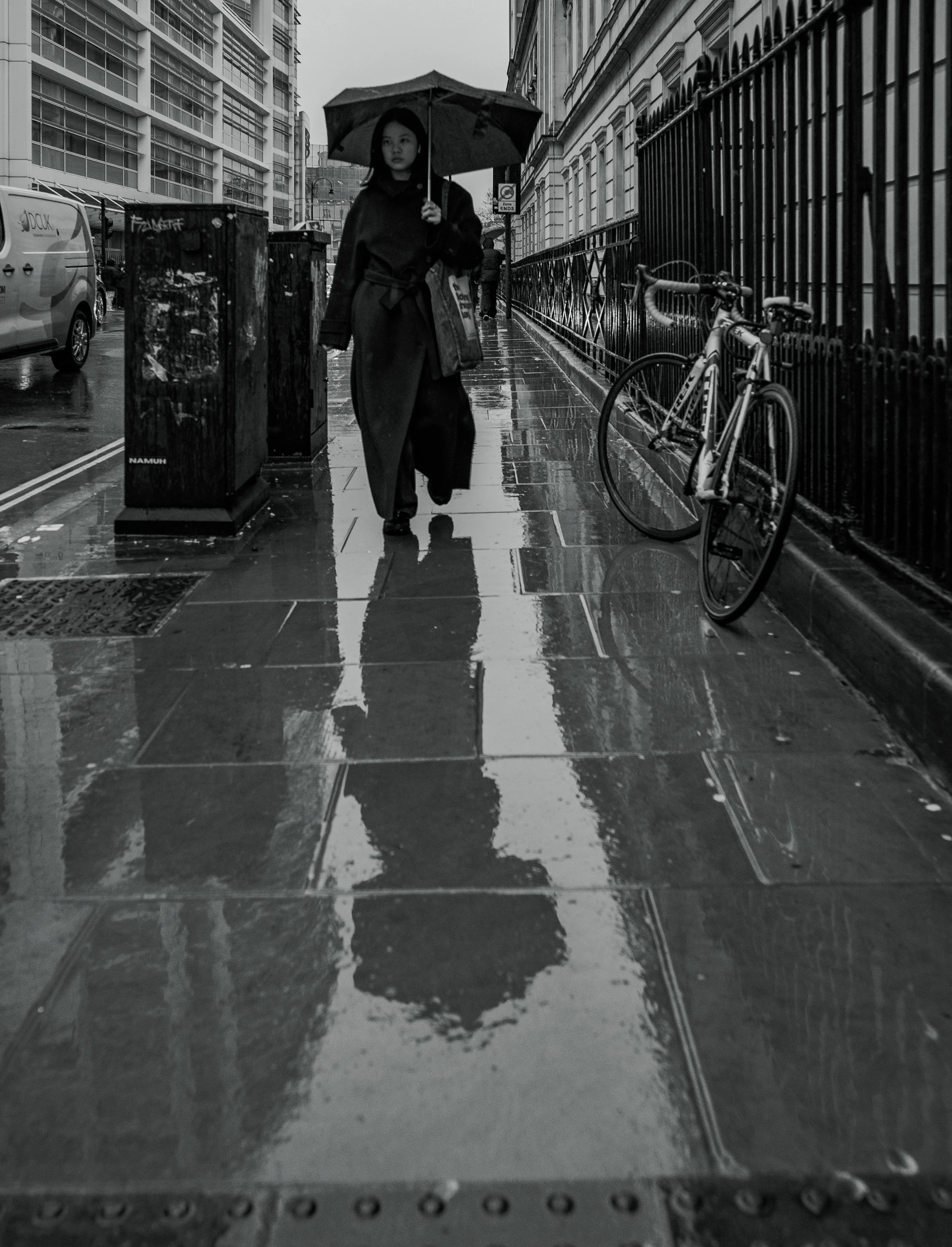 A woman walking on a wet city sidewalk holding an umbrella, with her reflection visible on the puddles, a bicycle leaning against a fence, and buildings and vehicles in the background.