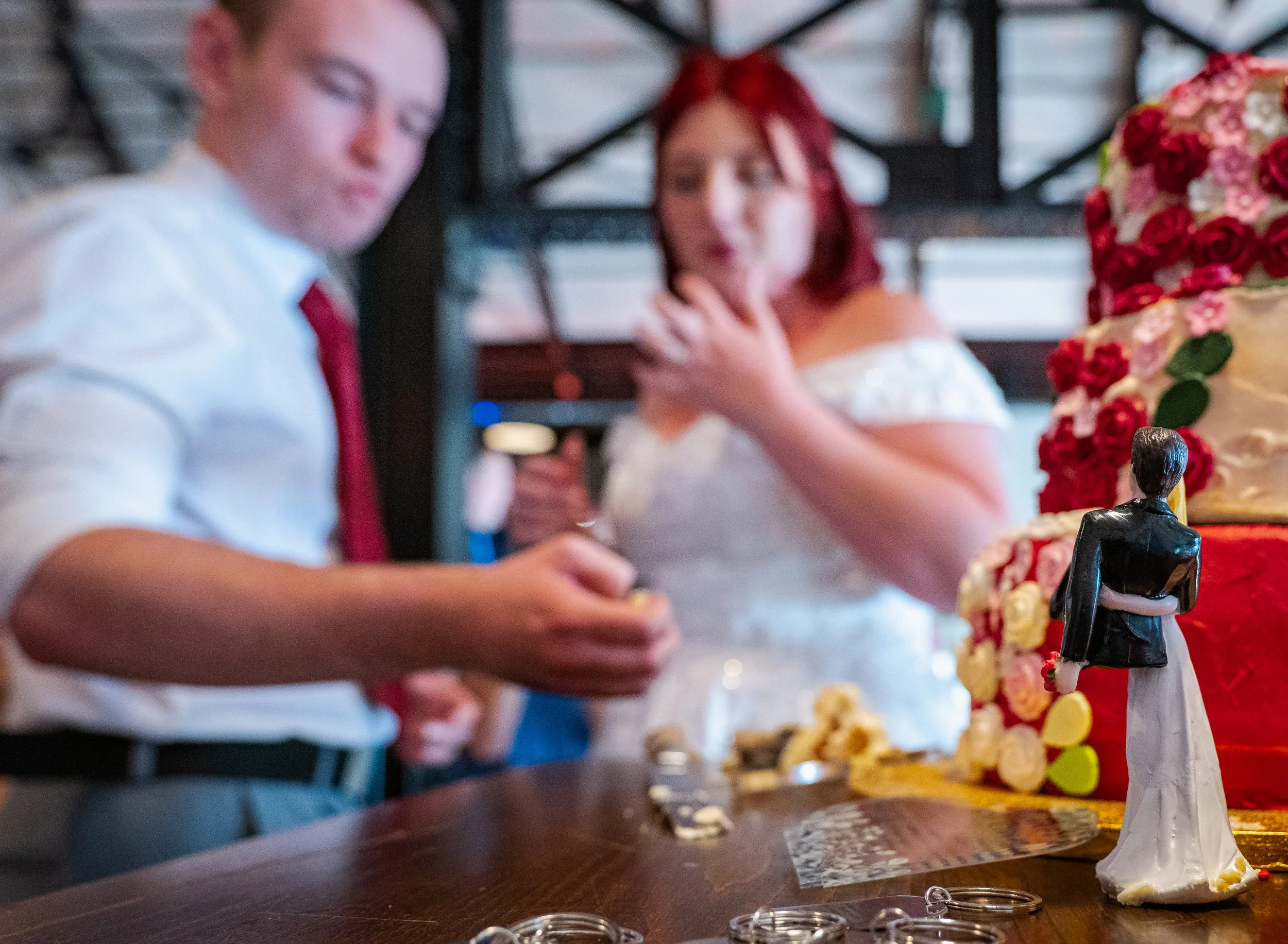 A wedding cake decorated with red, pink, and white flowers, with wedding cake toppers including a figurine of a bride and groom.