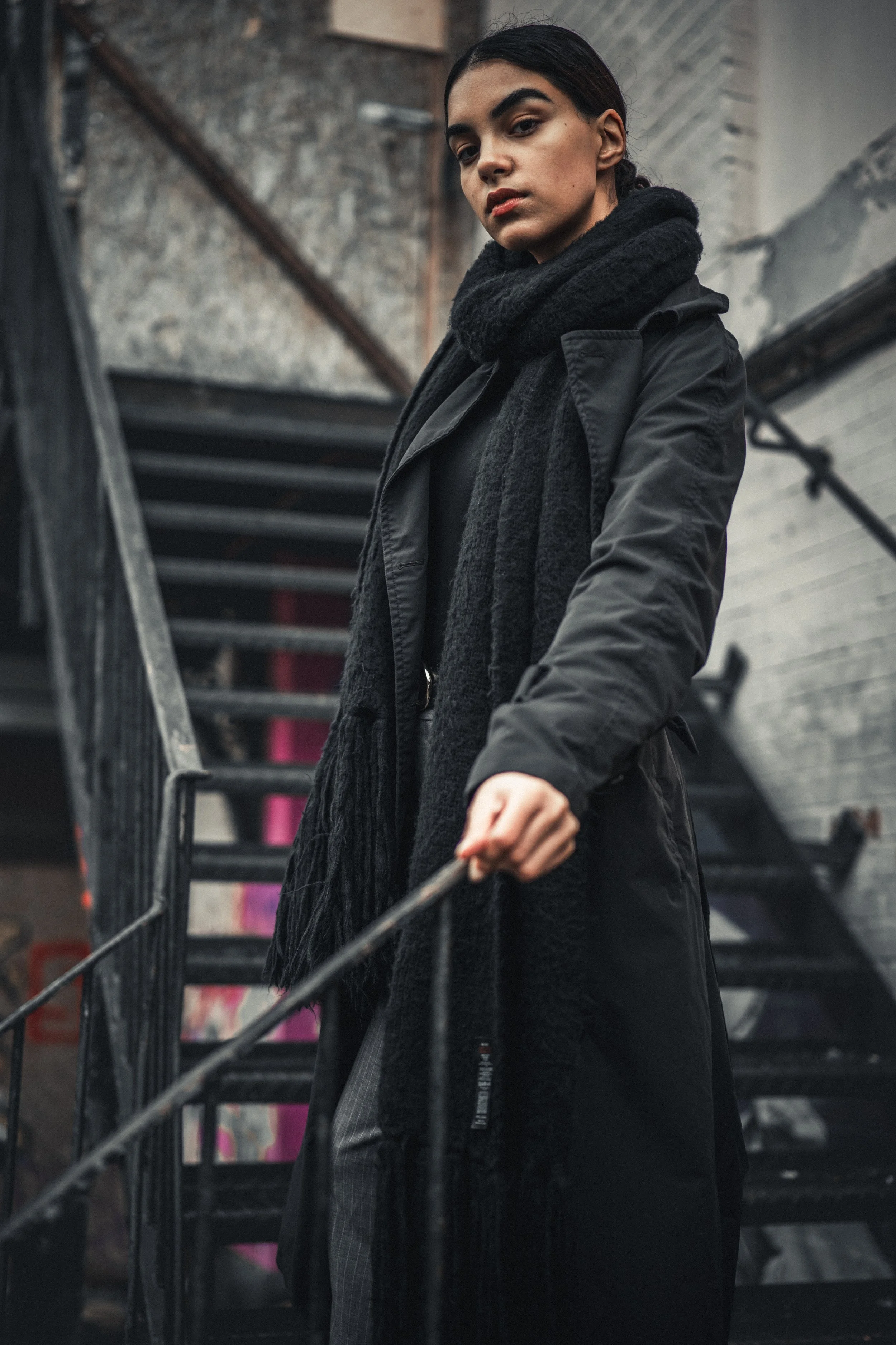 A woman standing on a metal staircase in an urban setting, wearing a black coat and scarf.