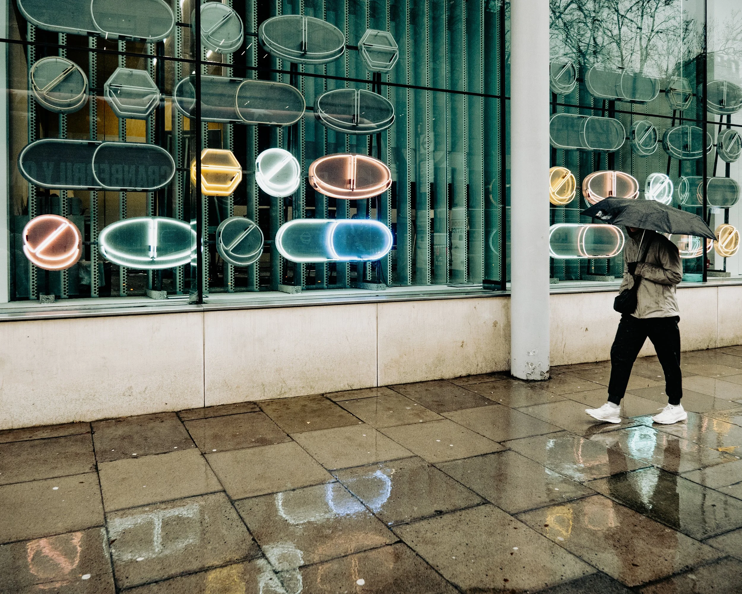 A person with a black umbrella walking on a wet sidewalk in front of a store window with neon signs and abstract shapes.