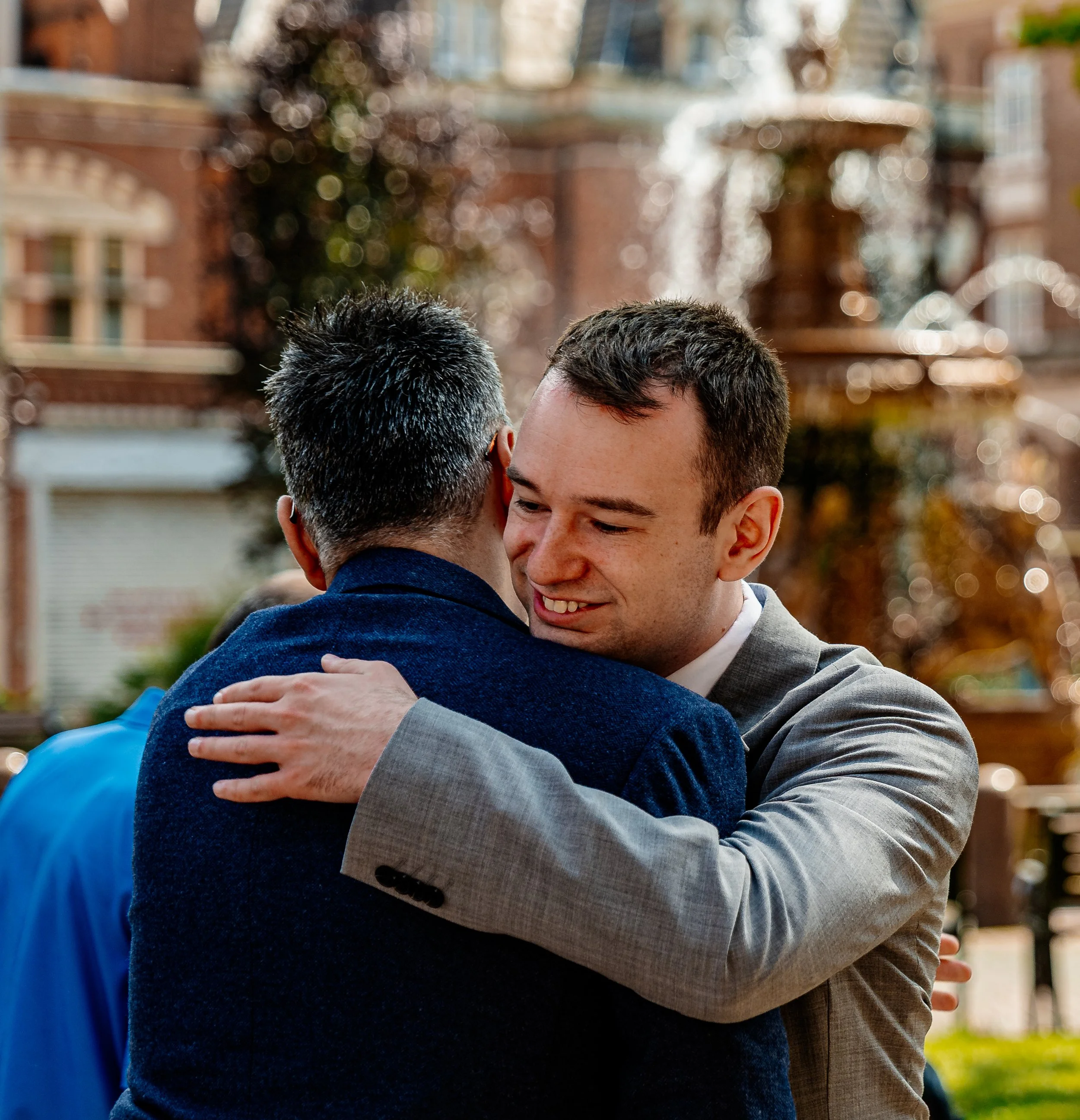Two men hugging outdoors, one with gray hair and wearing a blue jacket, the other with short dark hair and wearing a gray suit, smiling.