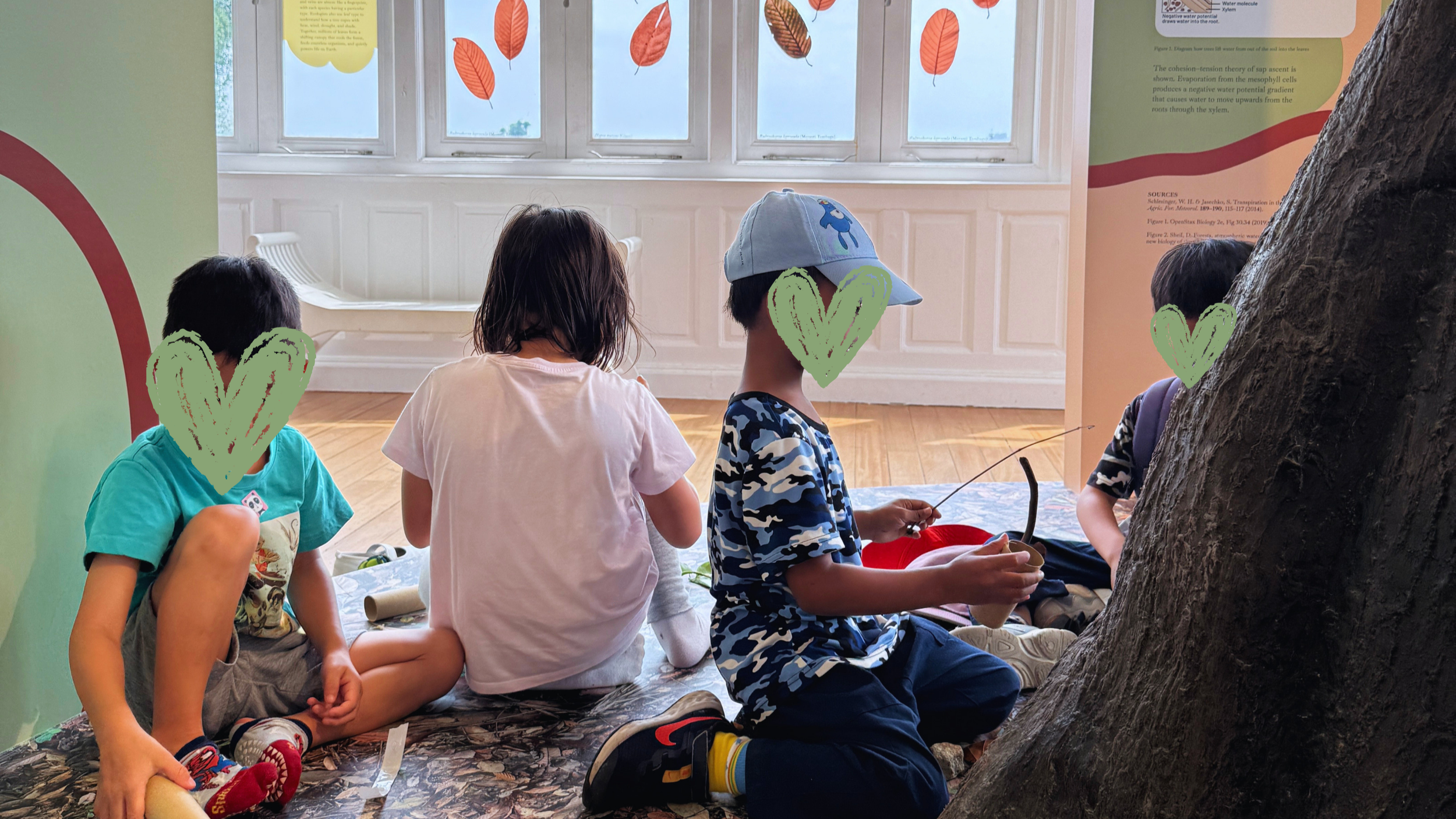 Children sitting on a display table at a museum, engaging with nature-themed exhibits, with leaf decorations on the window behind them.