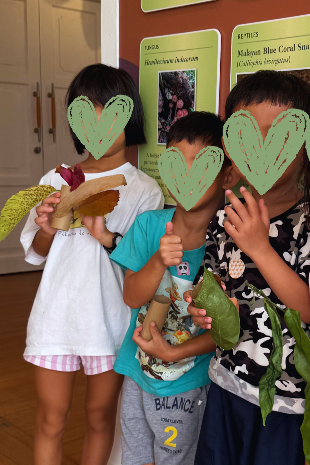 Three children at an educational exhibit holding plant and mushroom specimens, standing in front of informational posters about fungi and reptiles.