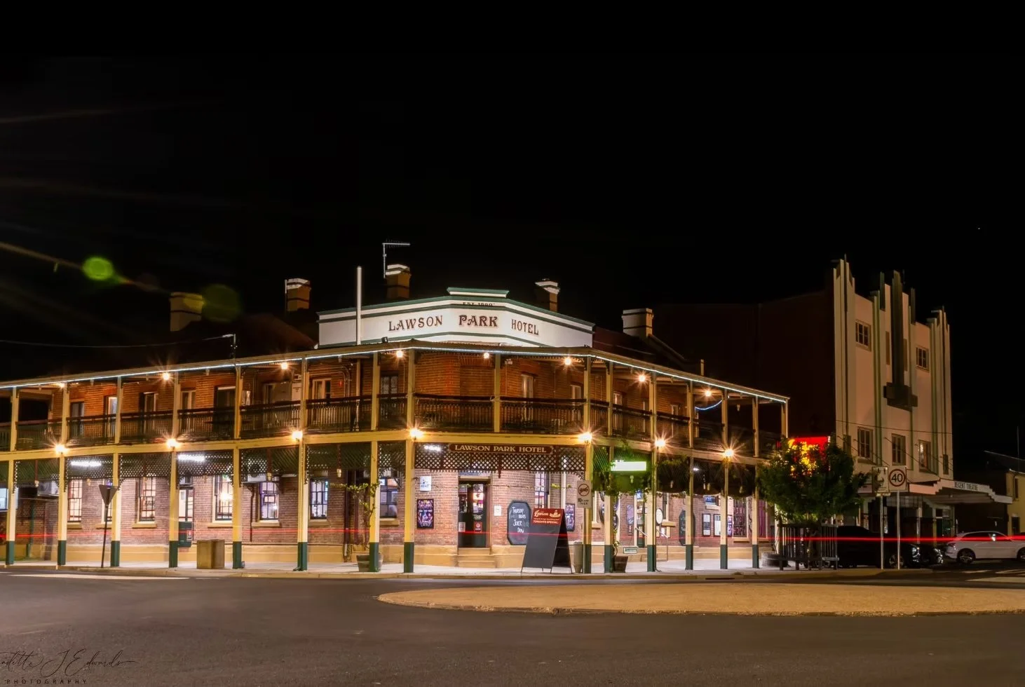 Night view of the historic Lawson Park Hotel, a two-story brick building with a wrap-around balcony adorned with string lights, and a sign on top displaying its name. The street in front is lit by streetlights, with parked cars and a few trees nearby.