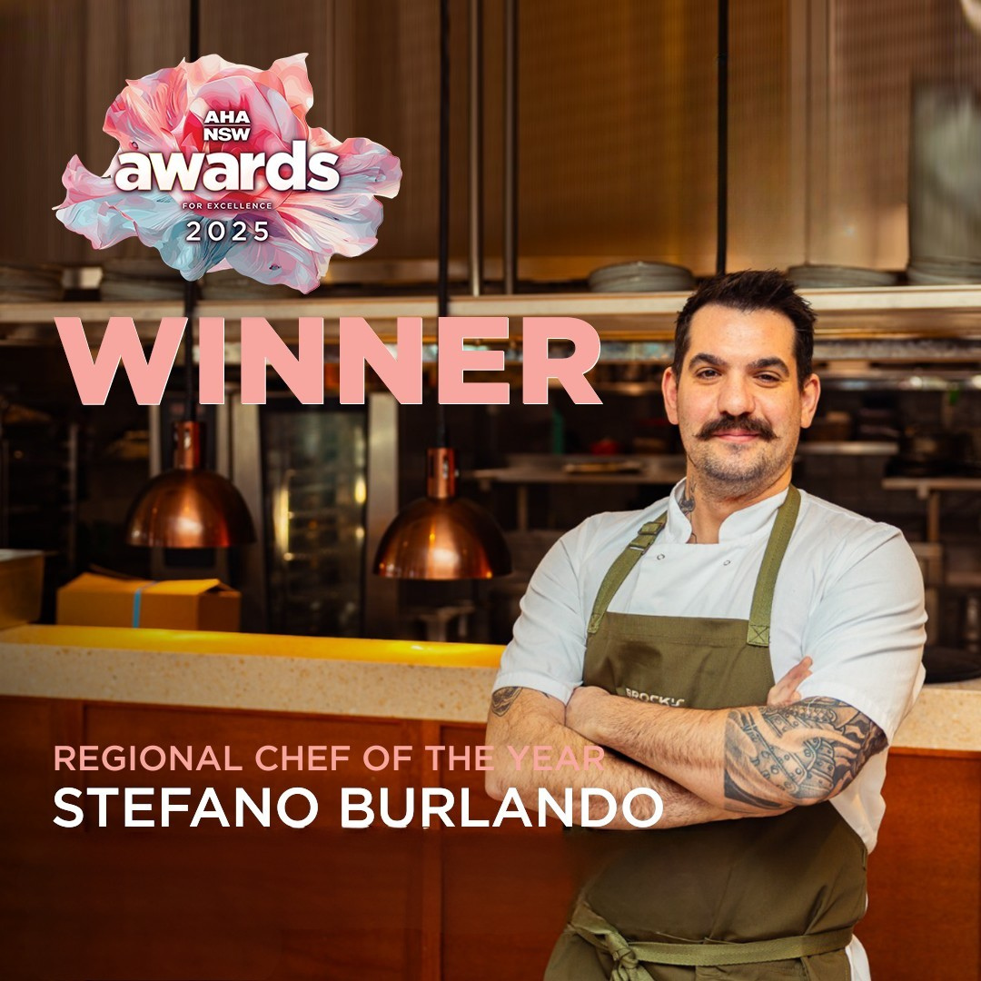 A man in a chef's uniform with tattoos on his arms, standing with arms crossed in a restaurant kitchen. Text overlay announces Stefano Burlando as the winner of the 2025 regional chef of the year award from AHA NSW.