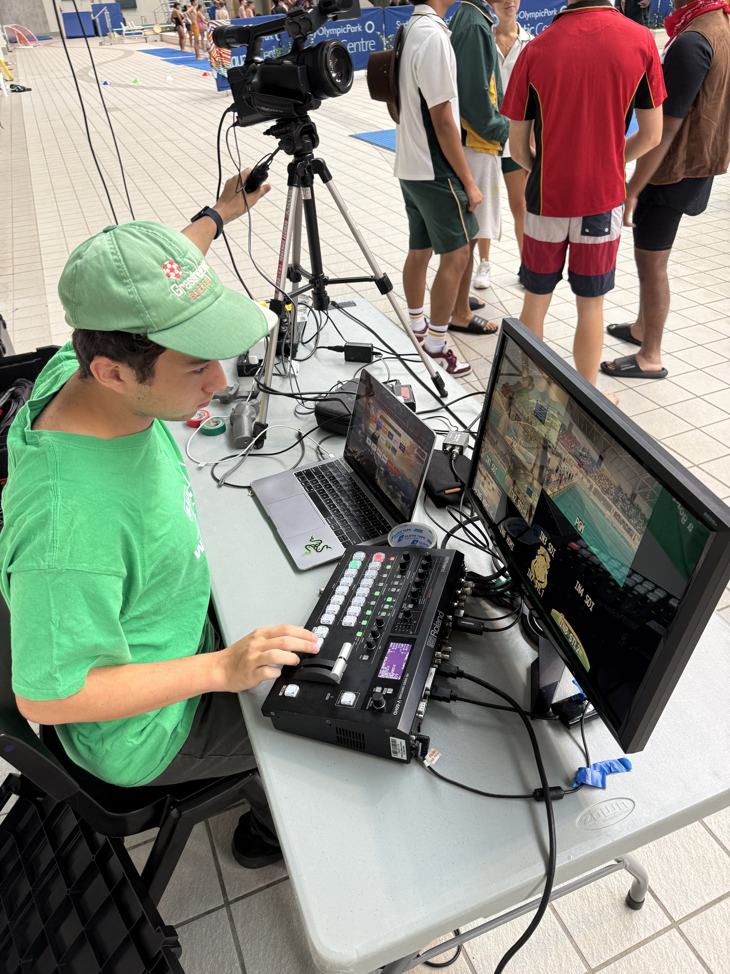 A technician sitting at a table operating a professional video or photo camera setup with multiple screens, a laptop, and audio equipment. Behind him, a group of people are standing in line at what appears to be a swimming pool or aquatic center with