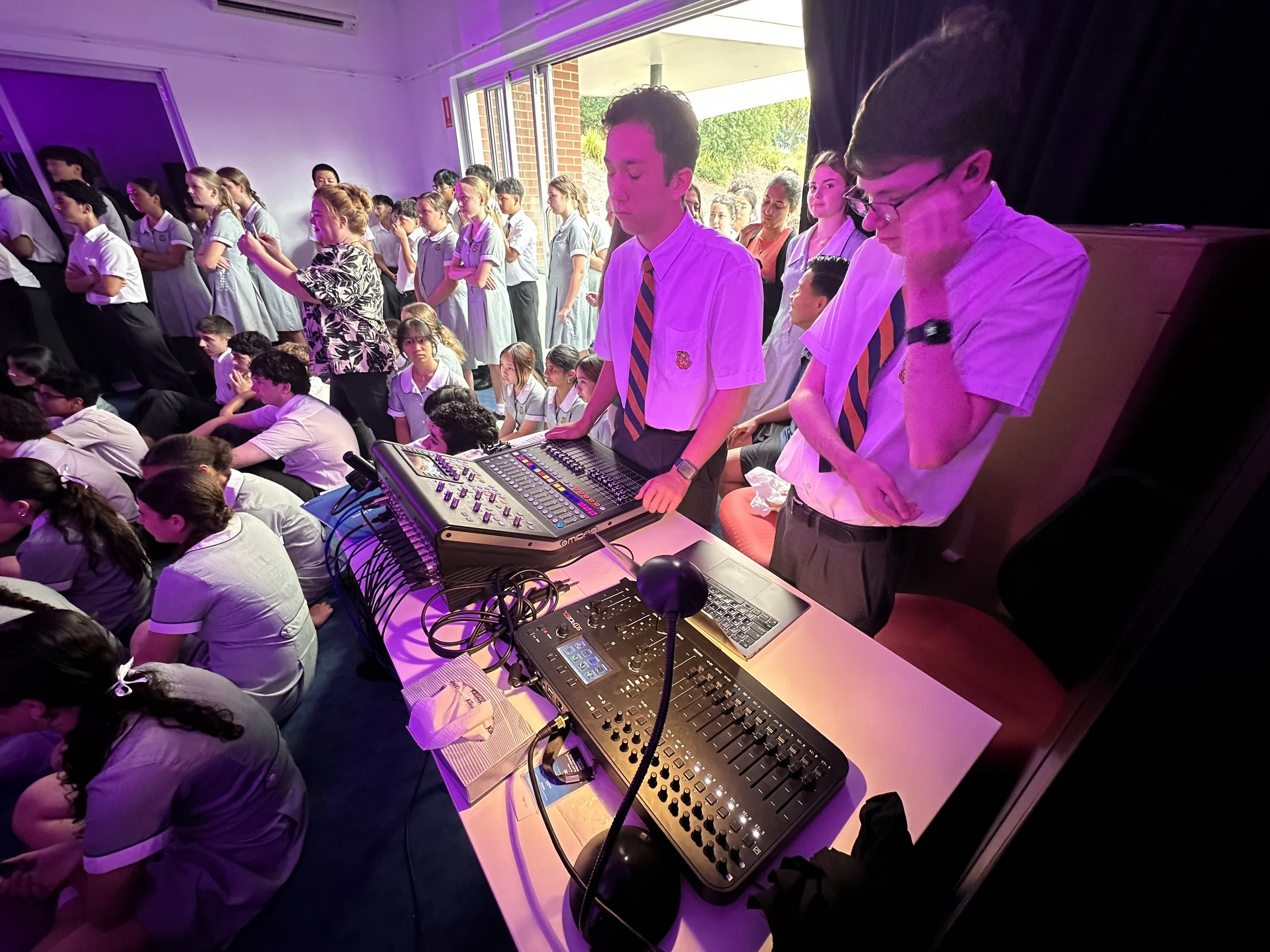 Students in school uniforms attending a school event, with some sitting on the floor and others standing, while a DJ plays music using professional sound equipment.