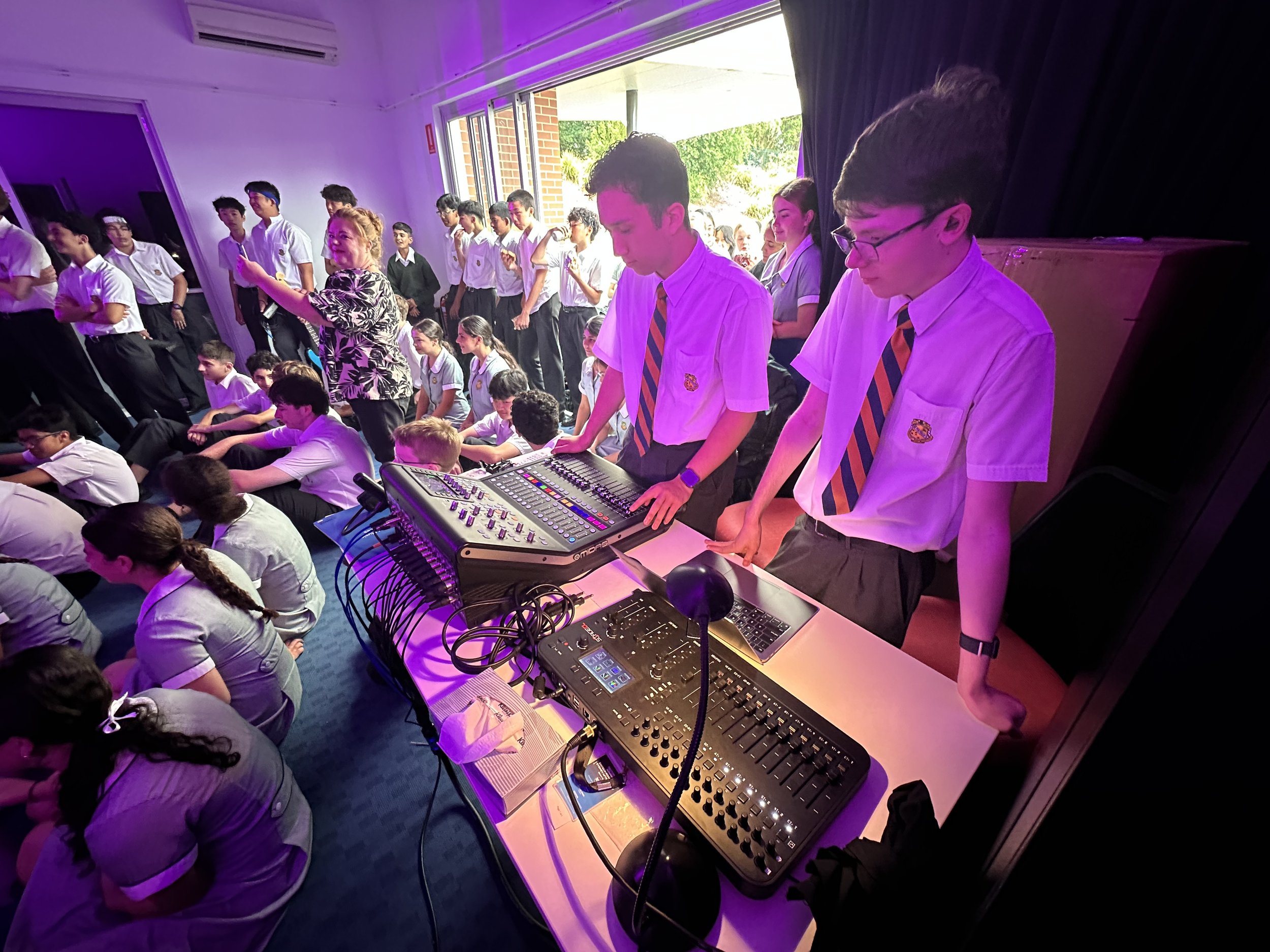 School assembly with students in uniform, some sitting on the floor and others standing, while two students operate audio equipment.