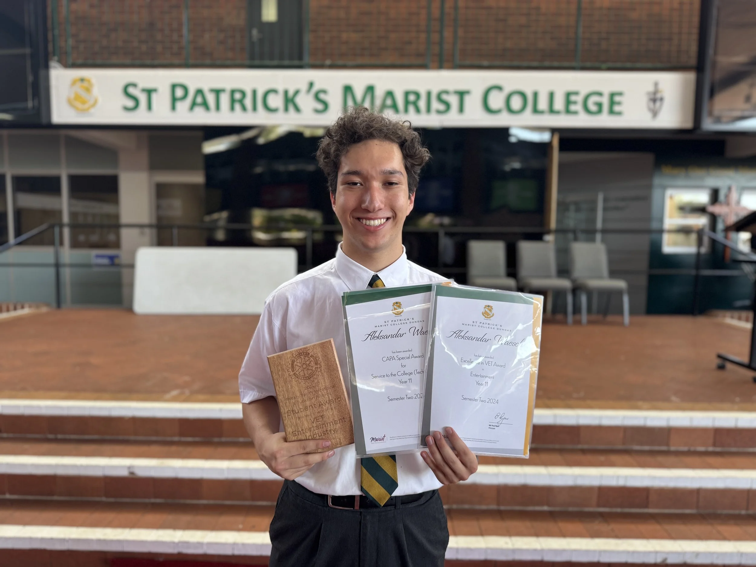 Young man in school uniform holding awards and certificate standing on steps in front of St. Patrick's Marist College sign.
