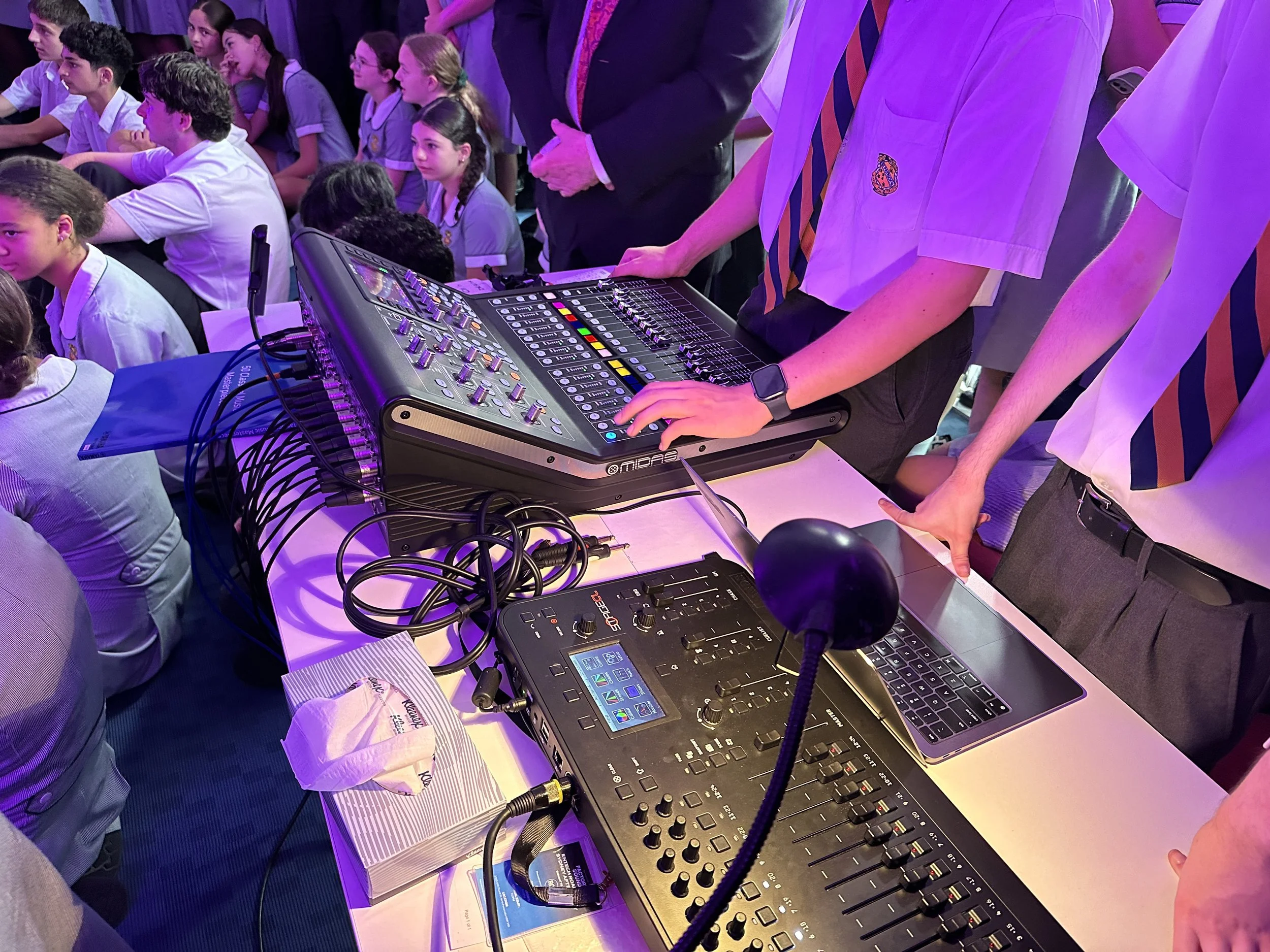 Students and adults in school uniforms gathered around a DJ station with sound equipment in a classroom or auditorium.