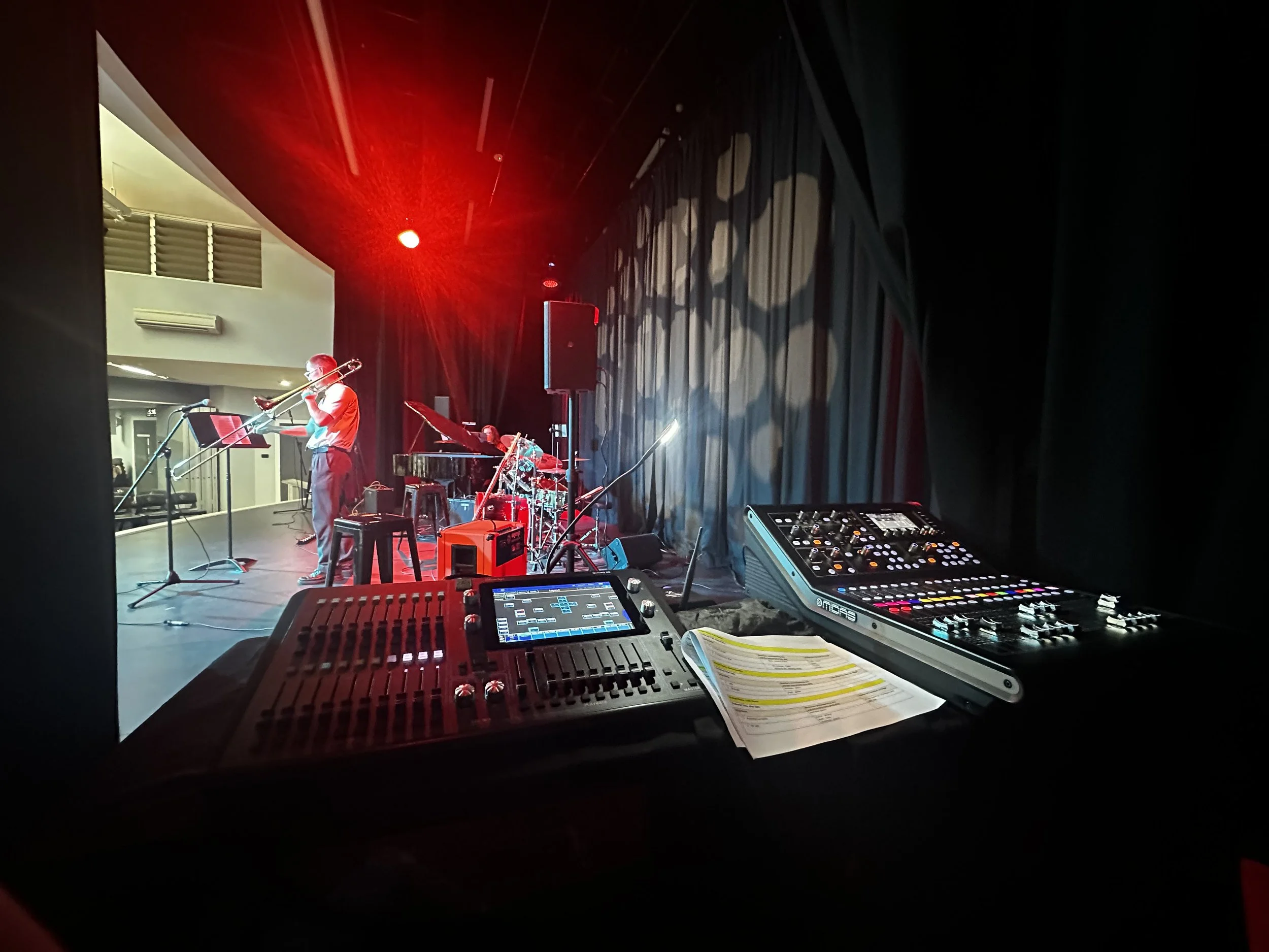 View from a sound booth overlooking a jazz band on stage, featuring a trombone player, a drummer, and a pianist, with stage lighting and sound equipment in the foreground.