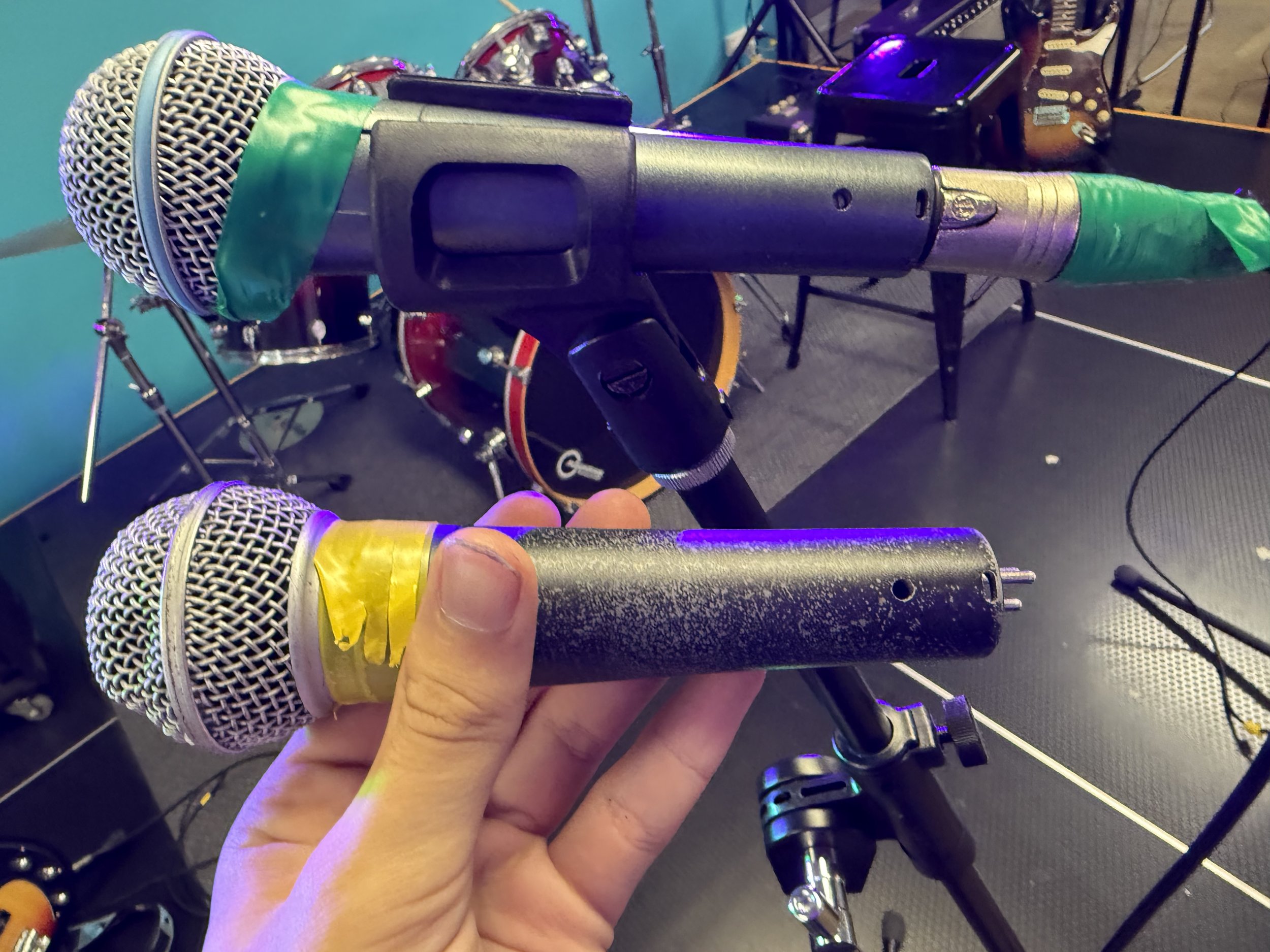 Two microphones, one with yellow tape near the grill and the other with green tape, held by a person's hand in a music or recording studio with drums and musical equipment in the background.