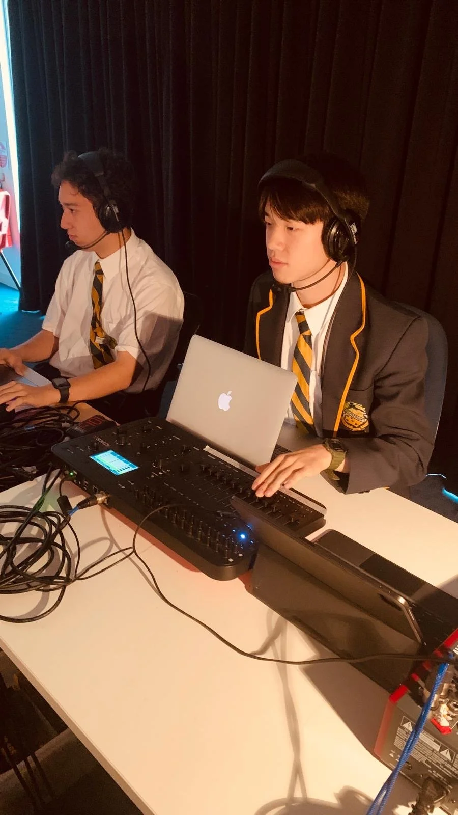 Two young men in school uniforms with yellow and black striped ties and blazers, wearing headphones, sitting at a table with audio equipment and laptops, participating in a sound or music production event.
