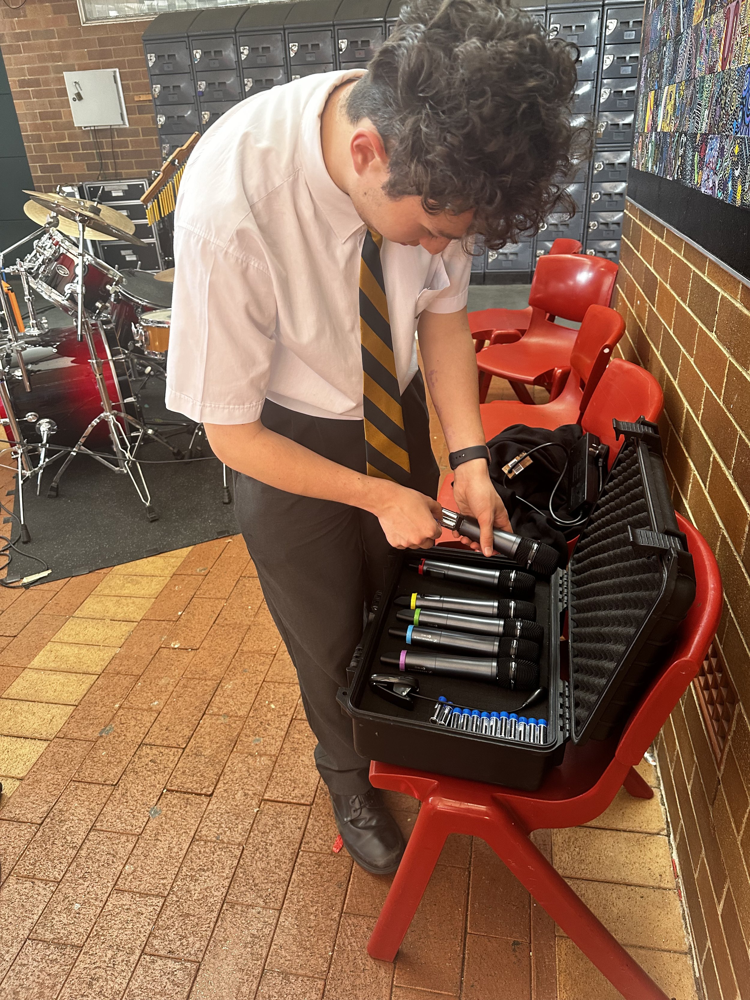 A young person in school uniform, including a white shirt and striped tie, standing next to a red bench, is organizing a set of microphones from a black carrying case. In the background, there is a drum set and some lockers.