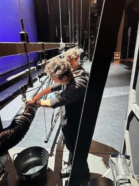 Two young men are working on a large stage or equipment rig, one is adjusting or inspecting a pipe or hose, with a bucket and tools nearby.