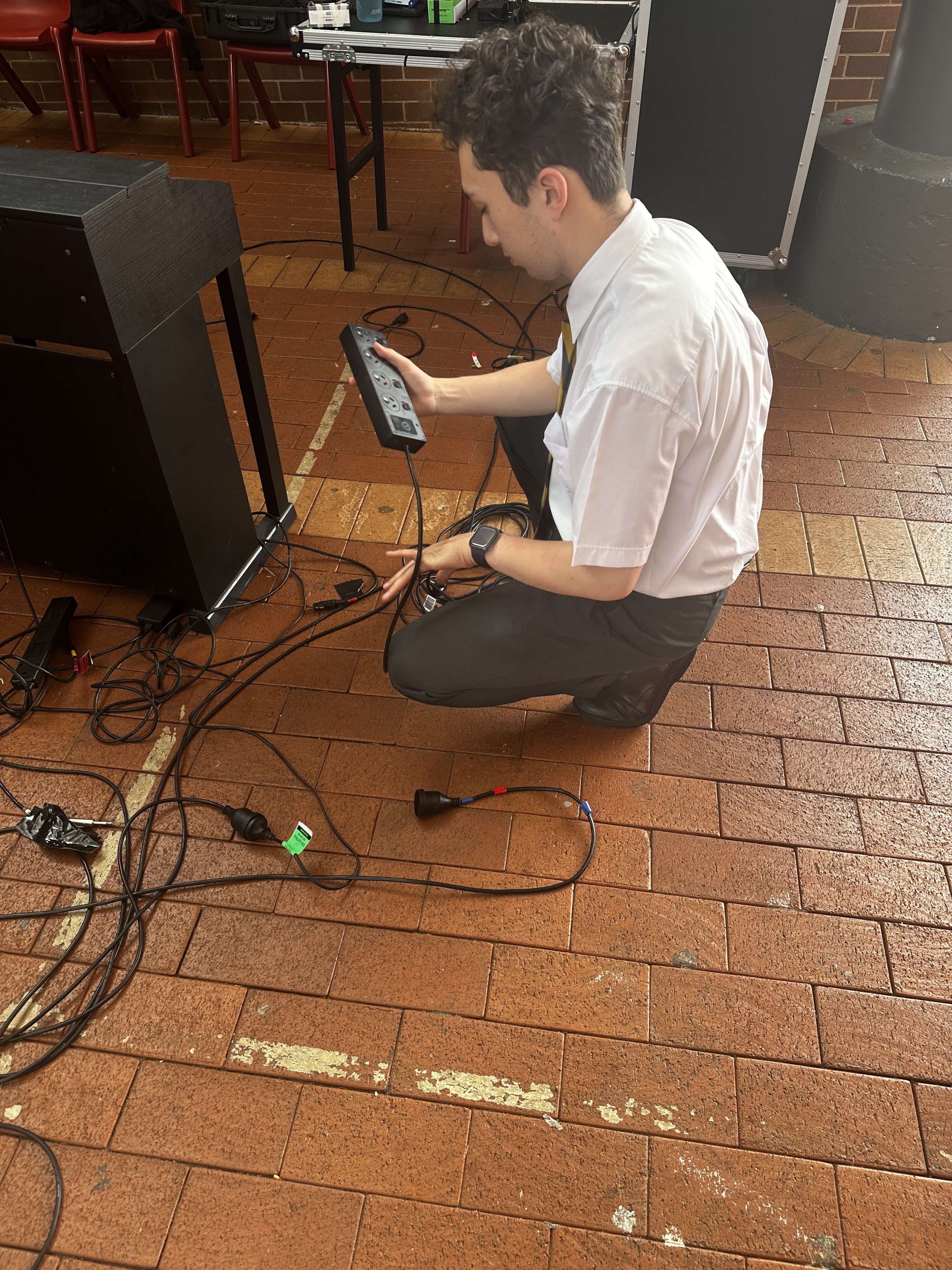 A young man in a white shirt and black pants kneels on a brick floor, holding a power strip with multiple outlets and plugged-in cables. There are tangled wires around him, and a black speaker or electronic device nearby. The background shows a brick