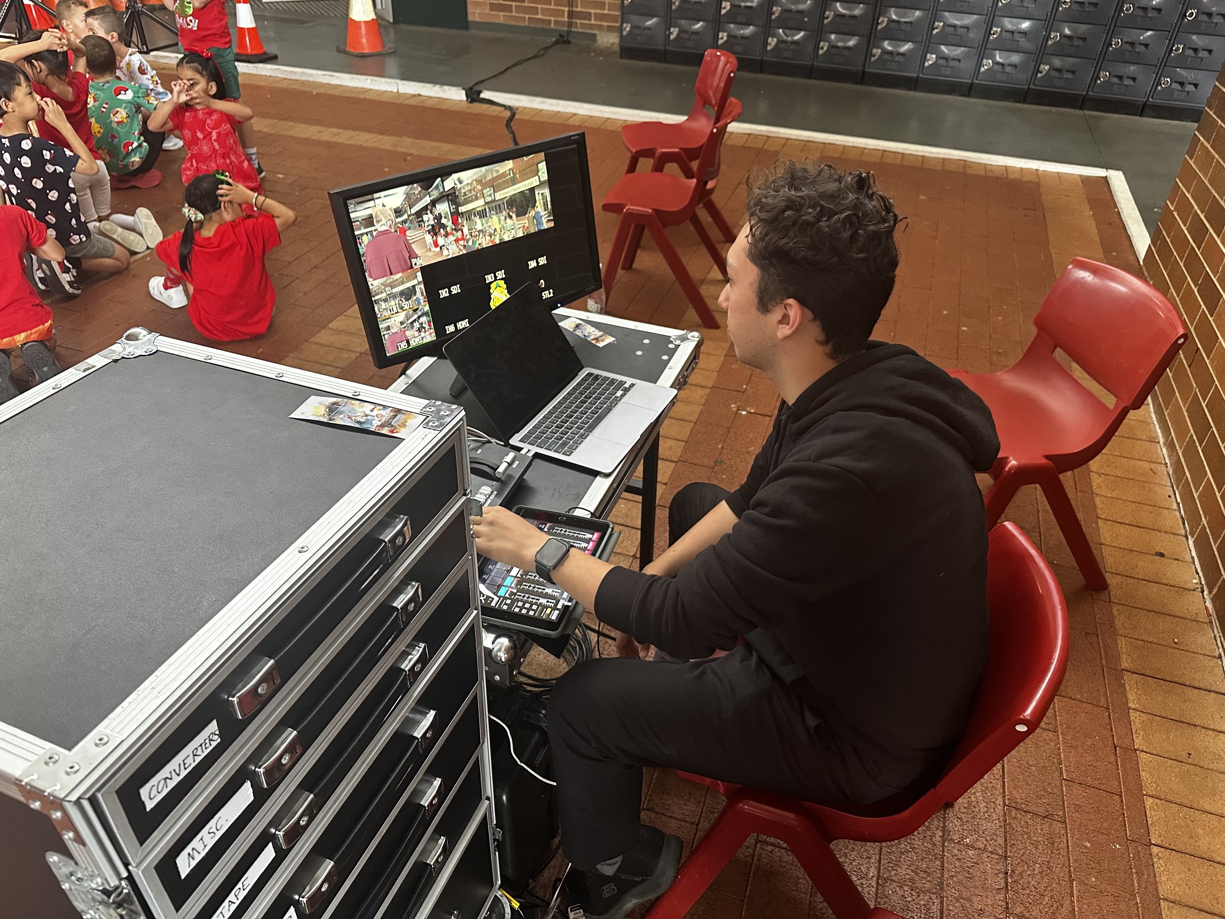 A man operating a video camera and laptop setup on a table, with kids dressed in festive clothing sitting on the floor nearby.