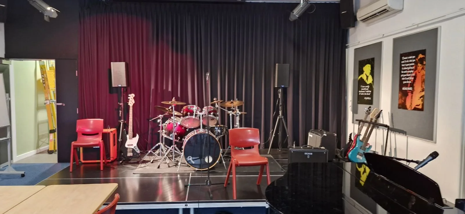 Music stage with drums, guitars, keyboard, microphones, and chairs, black curtains backdrop, framed posters on the side wall.