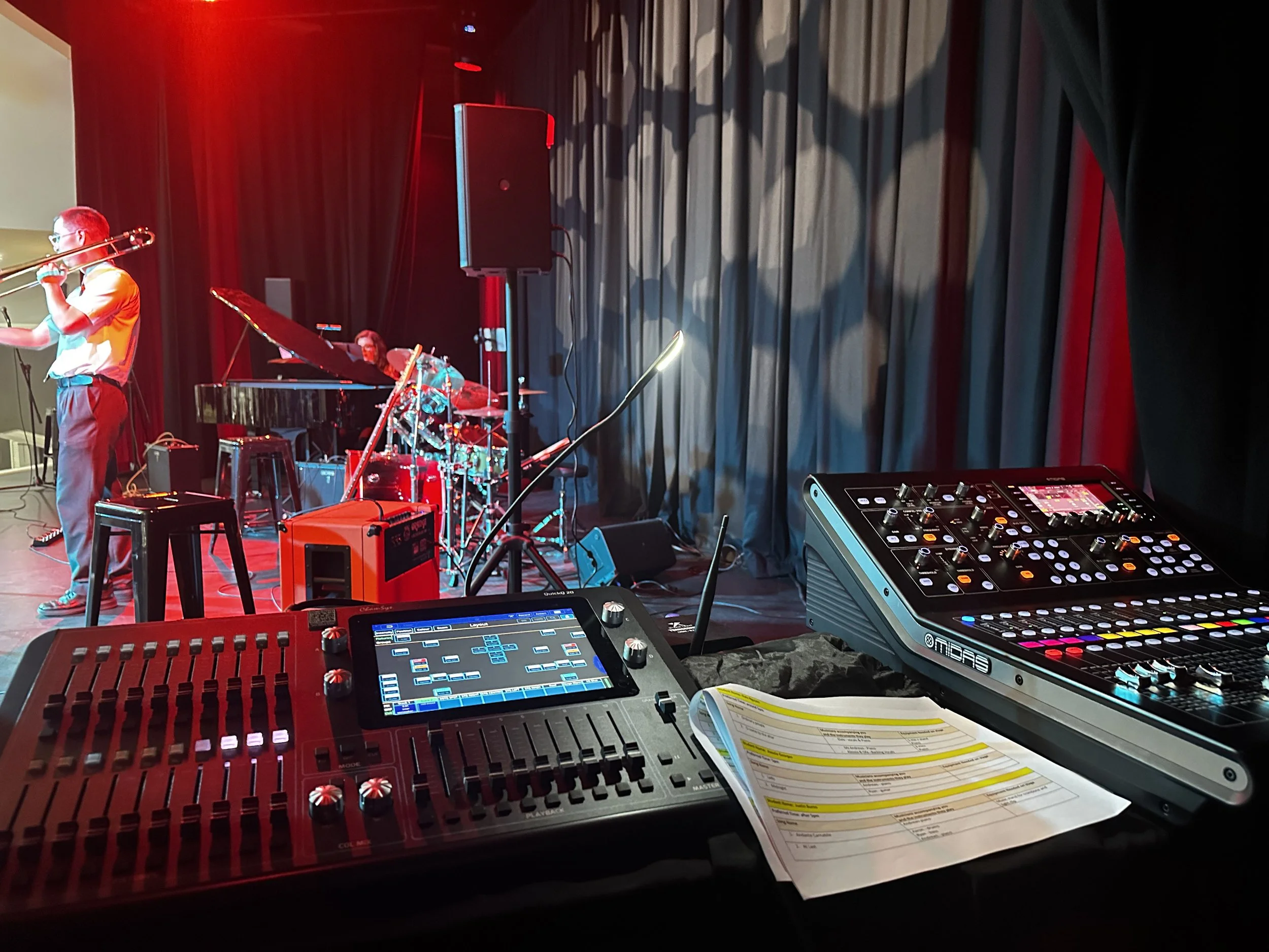 Concert stage with a musician playing the trombone, a drummer, and a grand piano in the background. Sound equipment and control panels in the foreground, with a black curtain backdrop.