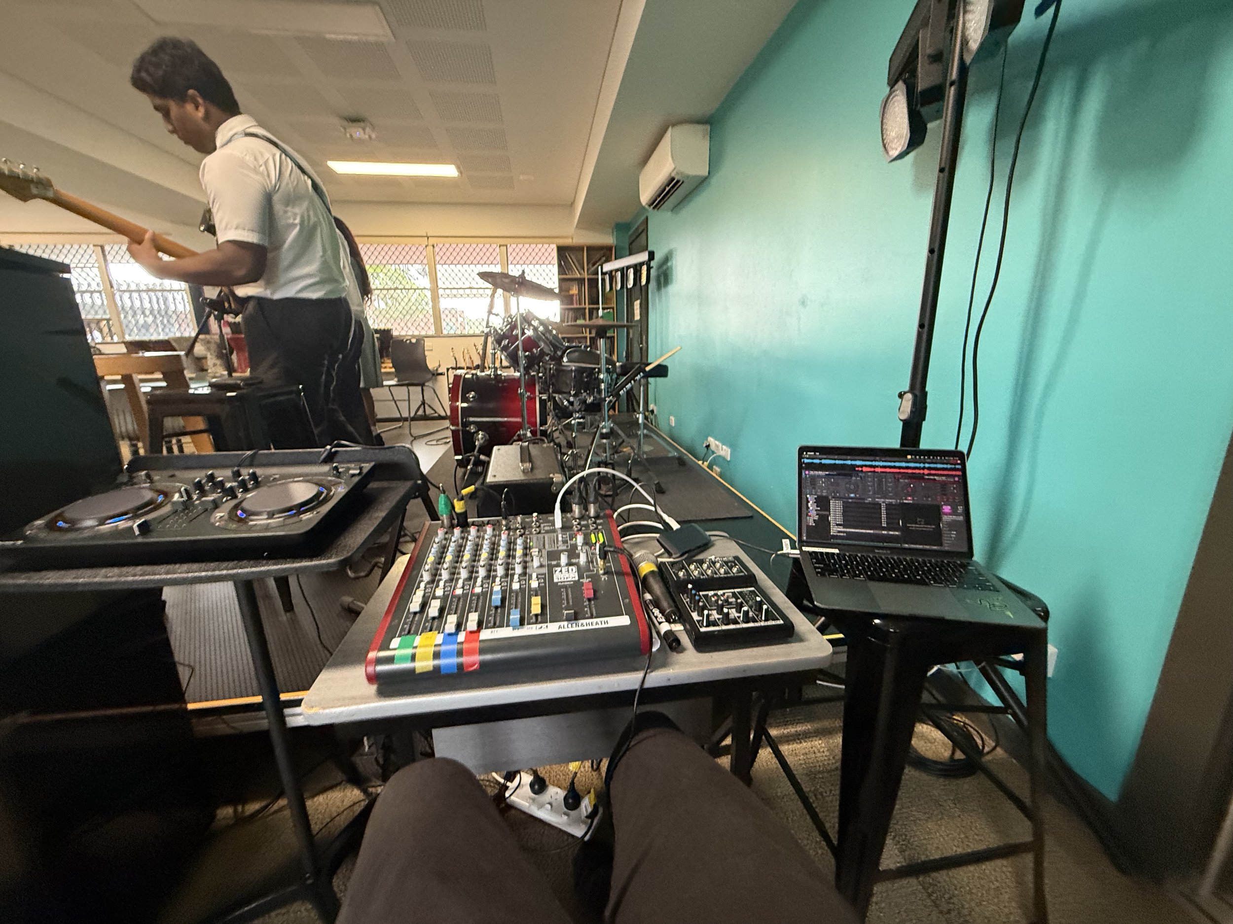 Music equipment including a DJ controller, mixer, laptop, and drums set up in a room with a teal wall and large window.