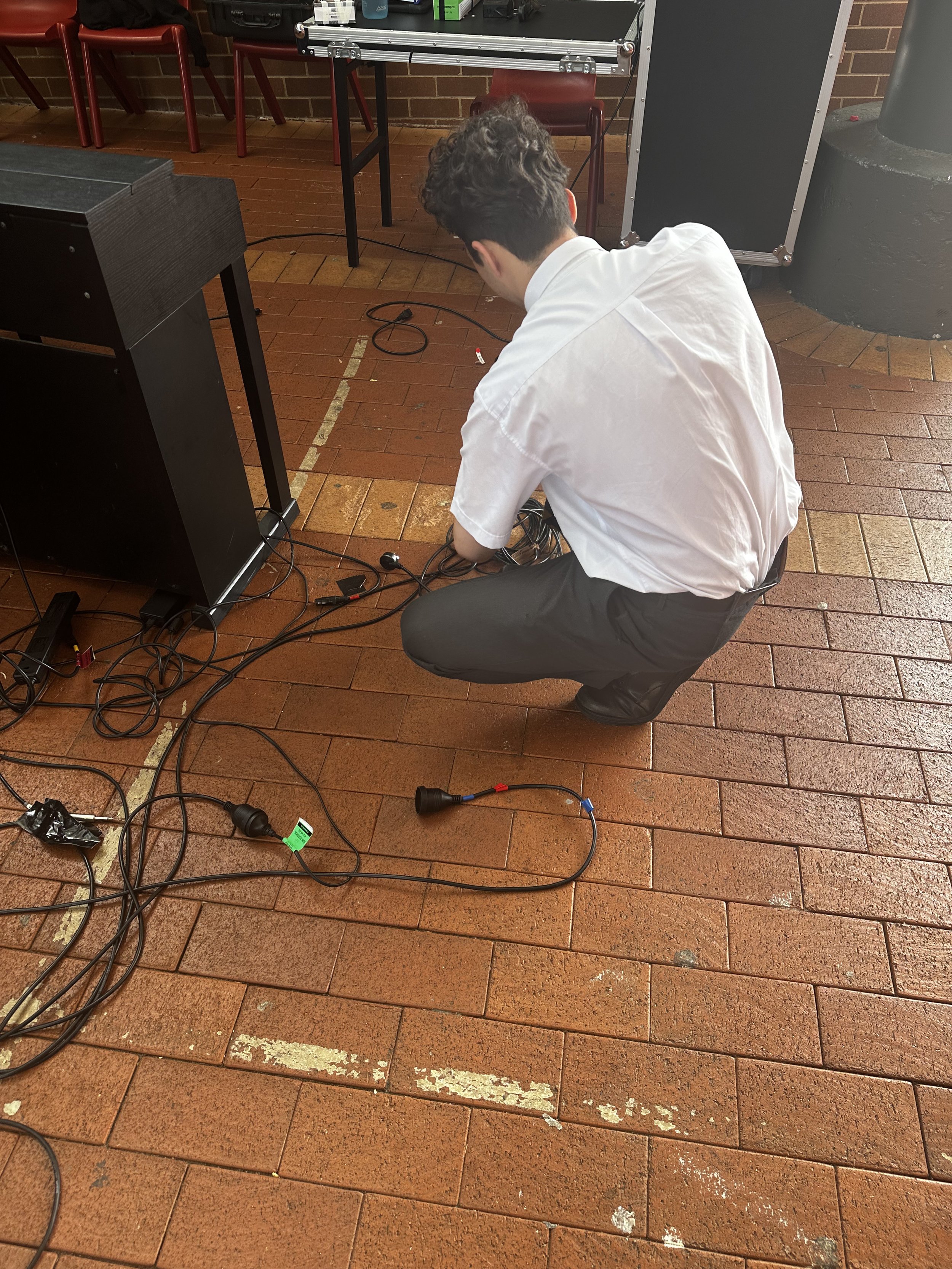 A man in a white shirt and black pants crouching on a brick floor, working with black cables and connectors near a table with electronic equipment.