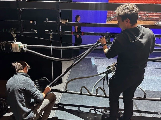 Two crew members operating camera and lighting equipment backstage in a theater, with a stage and audience seats in the background.