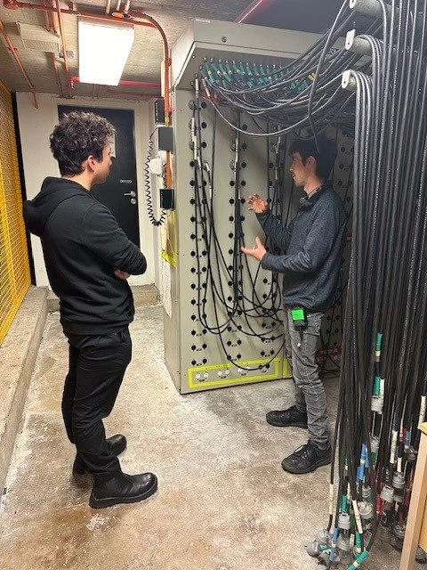 Two men stand in a utility room, engaged in conversation next to a large electrical panel with numerous cables.