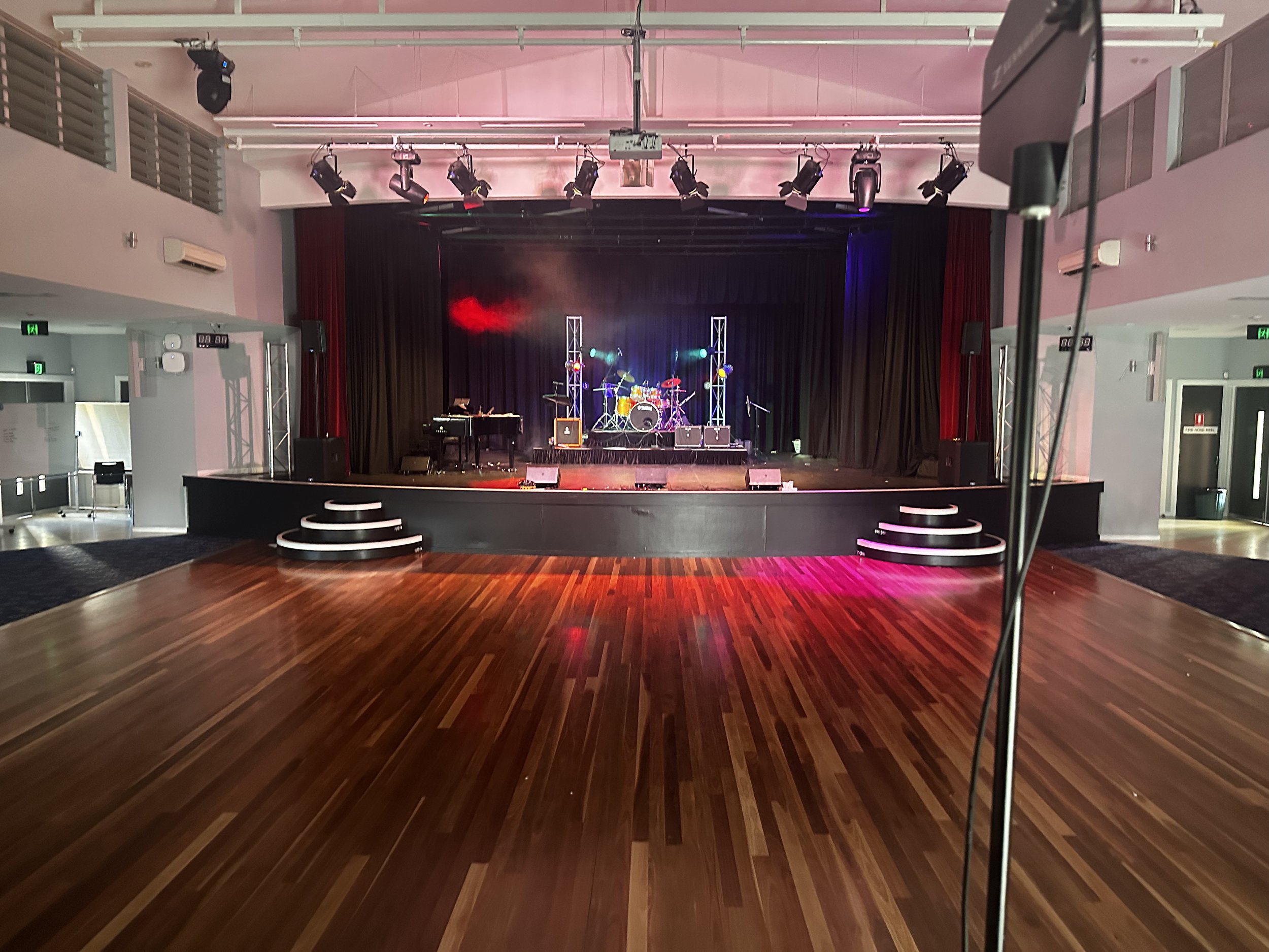 Empty stage with musical instruments, including a drum set, keyboard, microphone, and speakers, in a venue with overhead stage lighting and wooden dance floor.