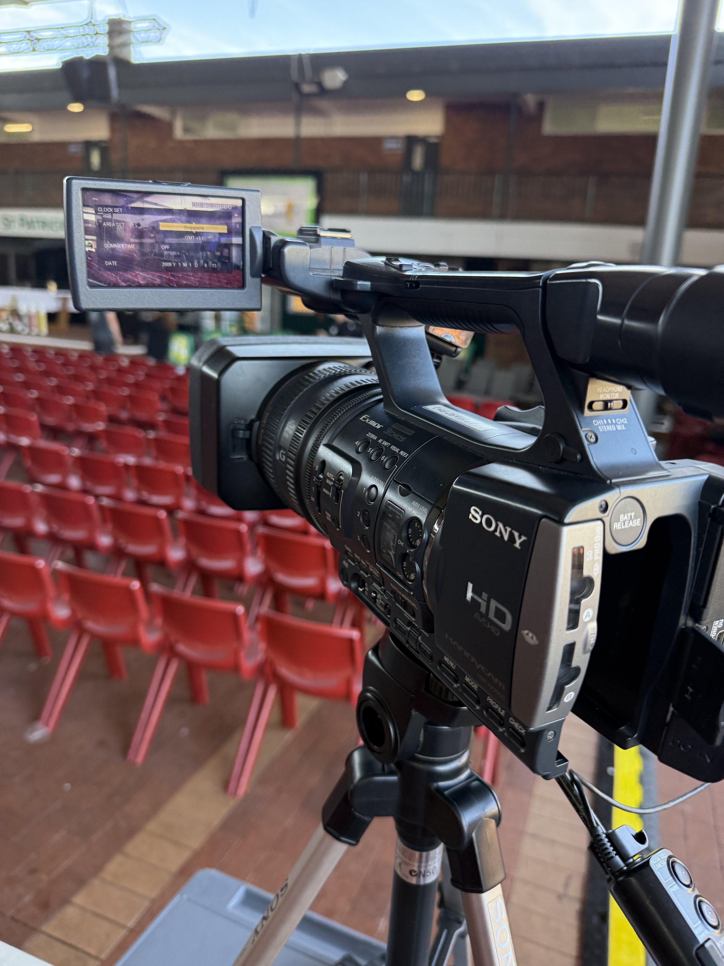 Close-up of a professional Sony HD camcorder mounted on a tripod, with empty red seats and a brick wall in the background, suggesting an event or concert venue.