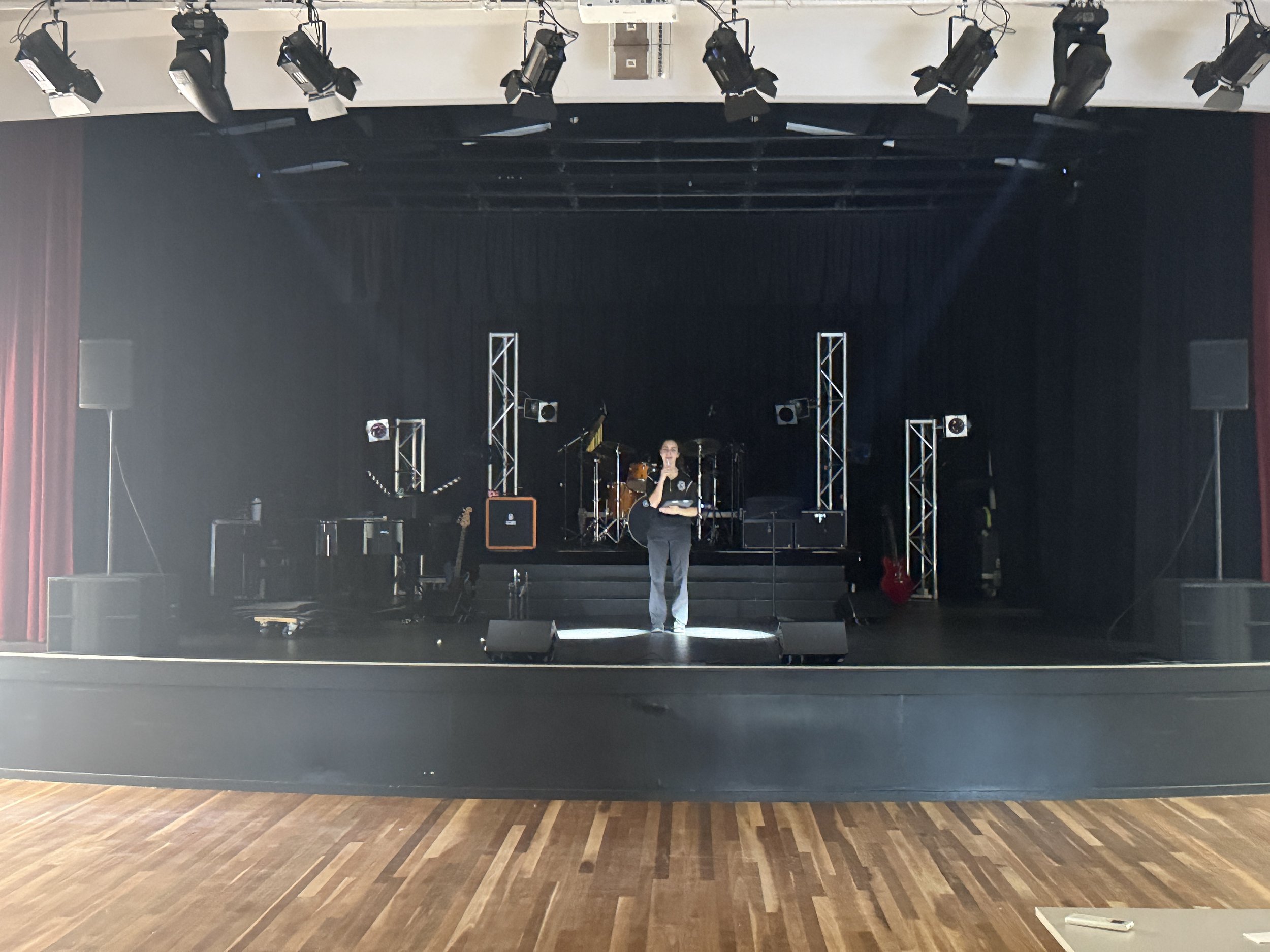 Empty stage with musical instruments and sound equipment, person standing center stage with microphone in hand.