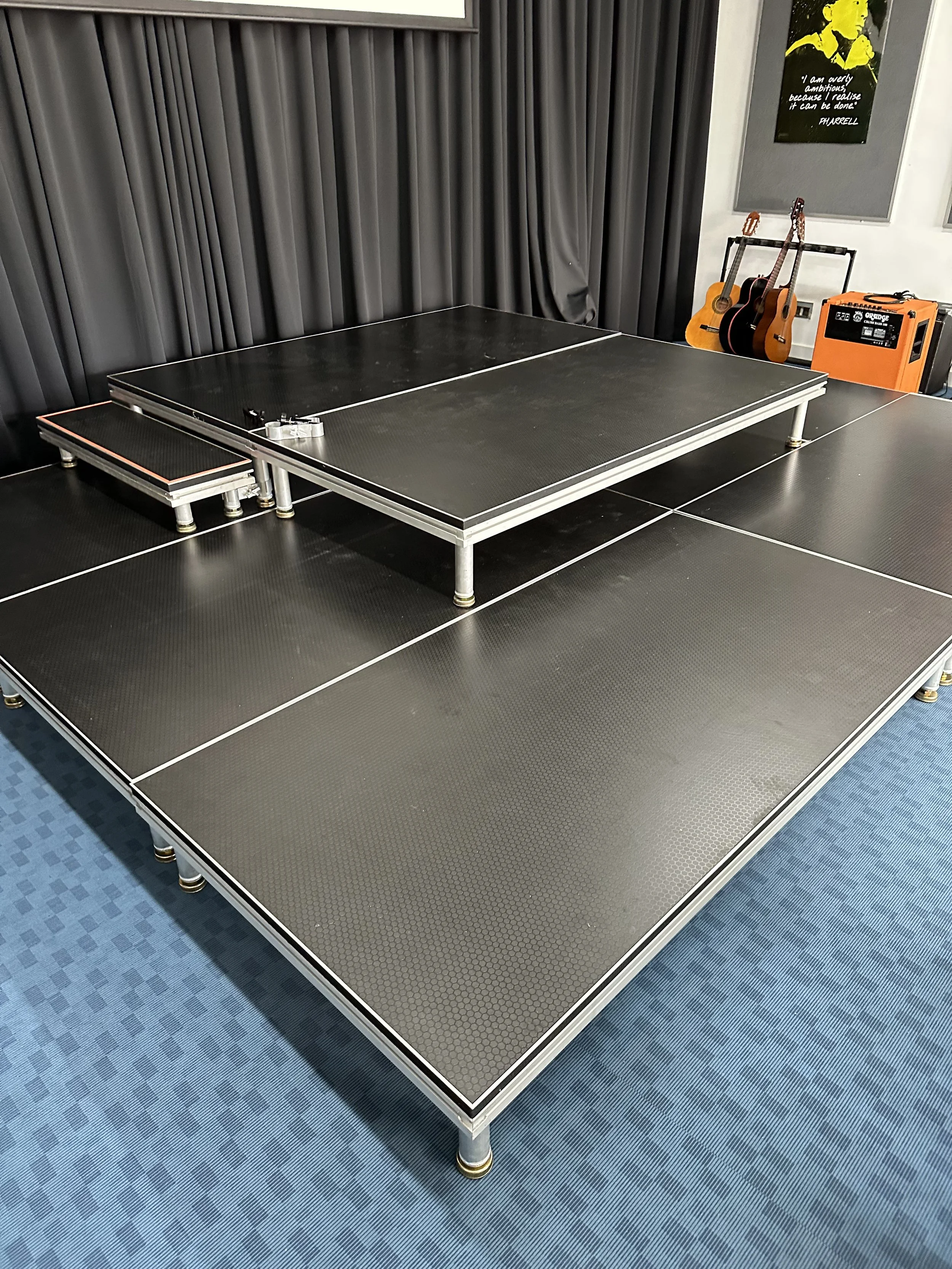 A small stage with a black surface, set up indoors with musical instruments and sound equipment in the background.