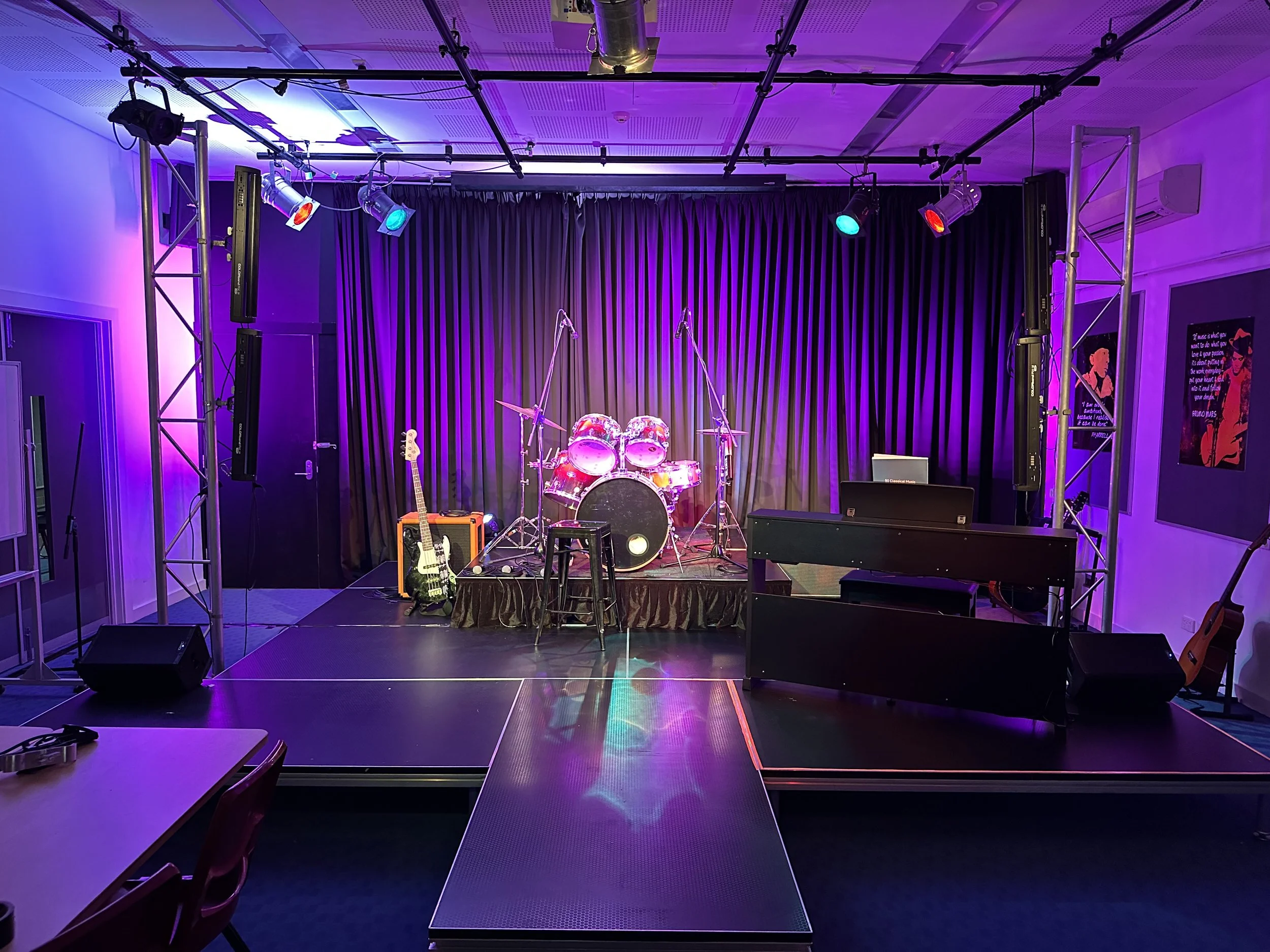 Empty stage set for a musical performance with purple lighting, a drum set, electric guitar, keyboard, and stage monitors.