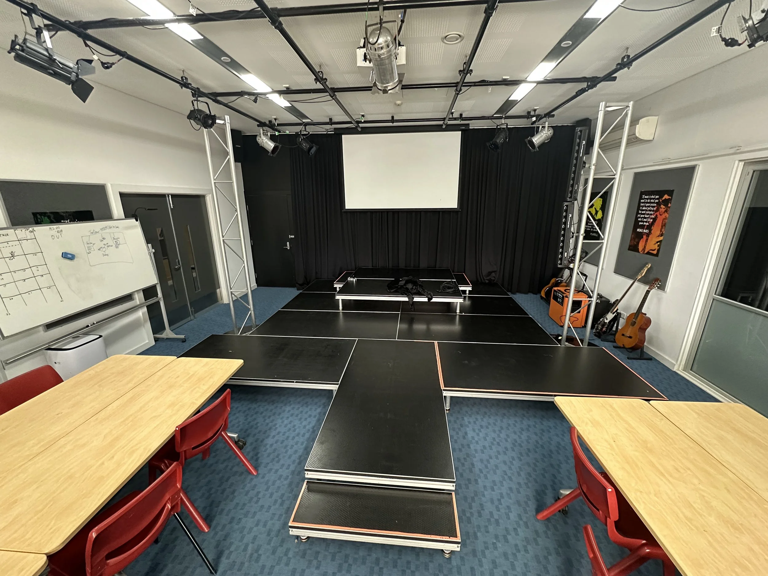 Empty stage with black curtains and giant white projector screen, two red chairs, folding tables, musical instruments including guitars, and a whiteboard with notes on it.