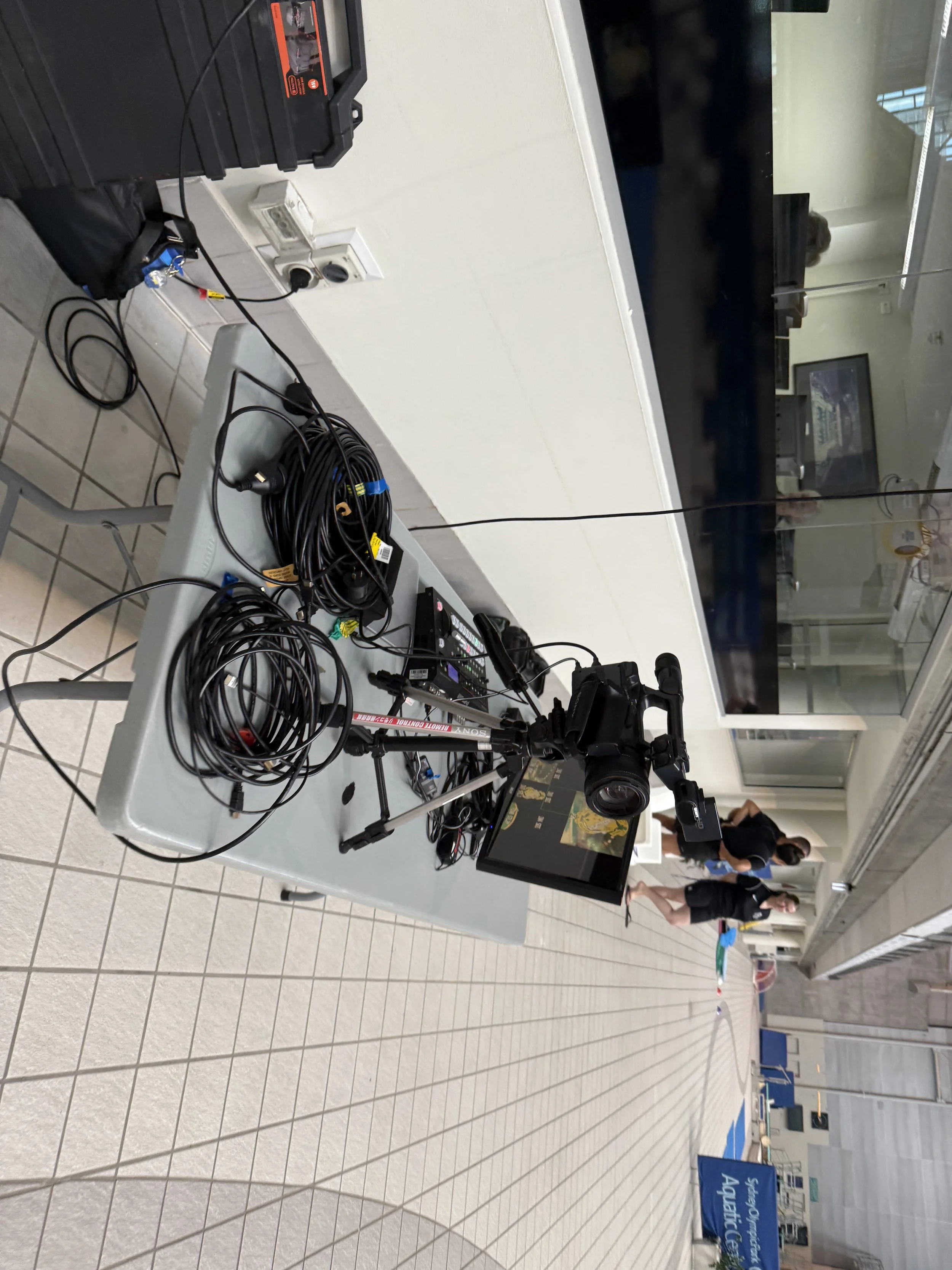 Equipment setup with cameras, cables, and monitors on a table in an indoor area, possibly for filming or broadcasting.