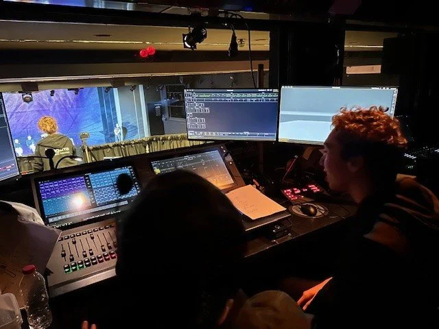 Behind-the-scenes view of a television or theater production control room with sound and video equipment, showing two people working at a console with multiple monitors, and a stage visible through a window in the background.