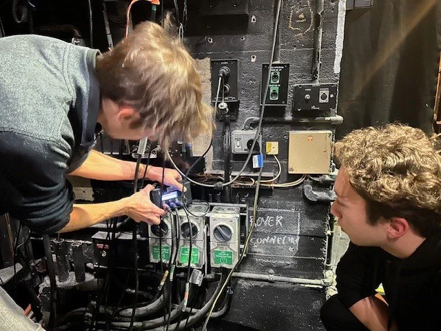 Two young men working on an electrical or technical setup with wires, switches, and control boxes on a black wall.