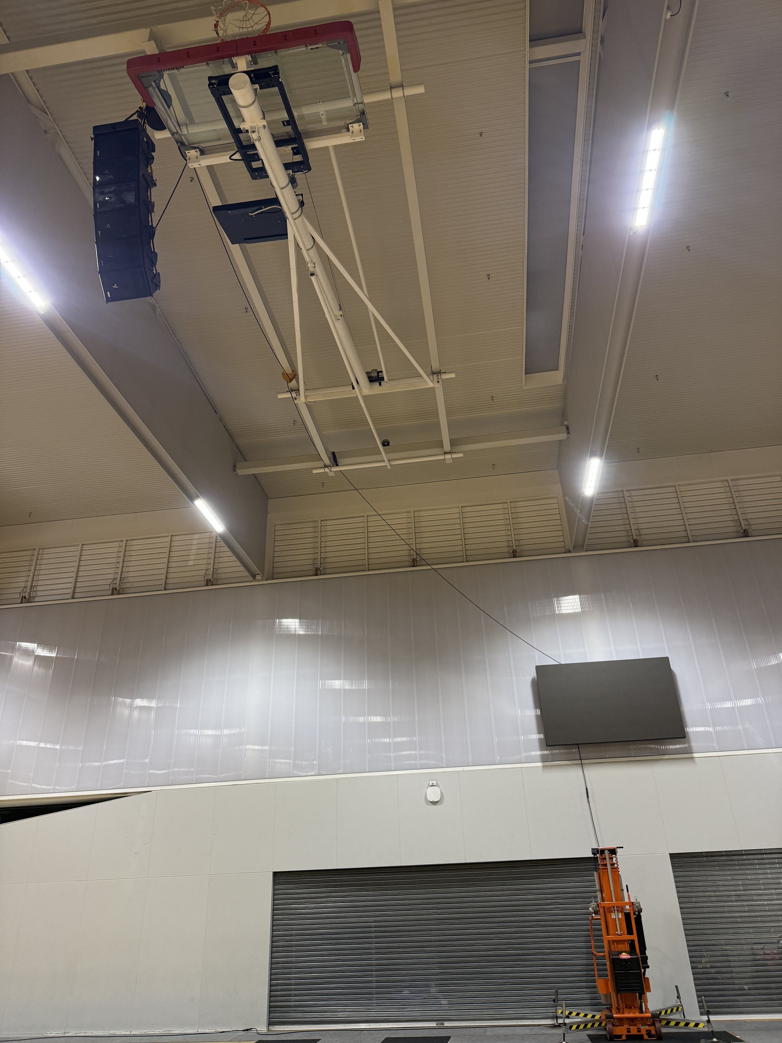 Indoor sports arena with a basketball hoop mounted on the ceiling and a large electronic scoreboard on the wall. Folded metal shutters cover windows or entrances, and an orange scissor lift is parked on the floor.