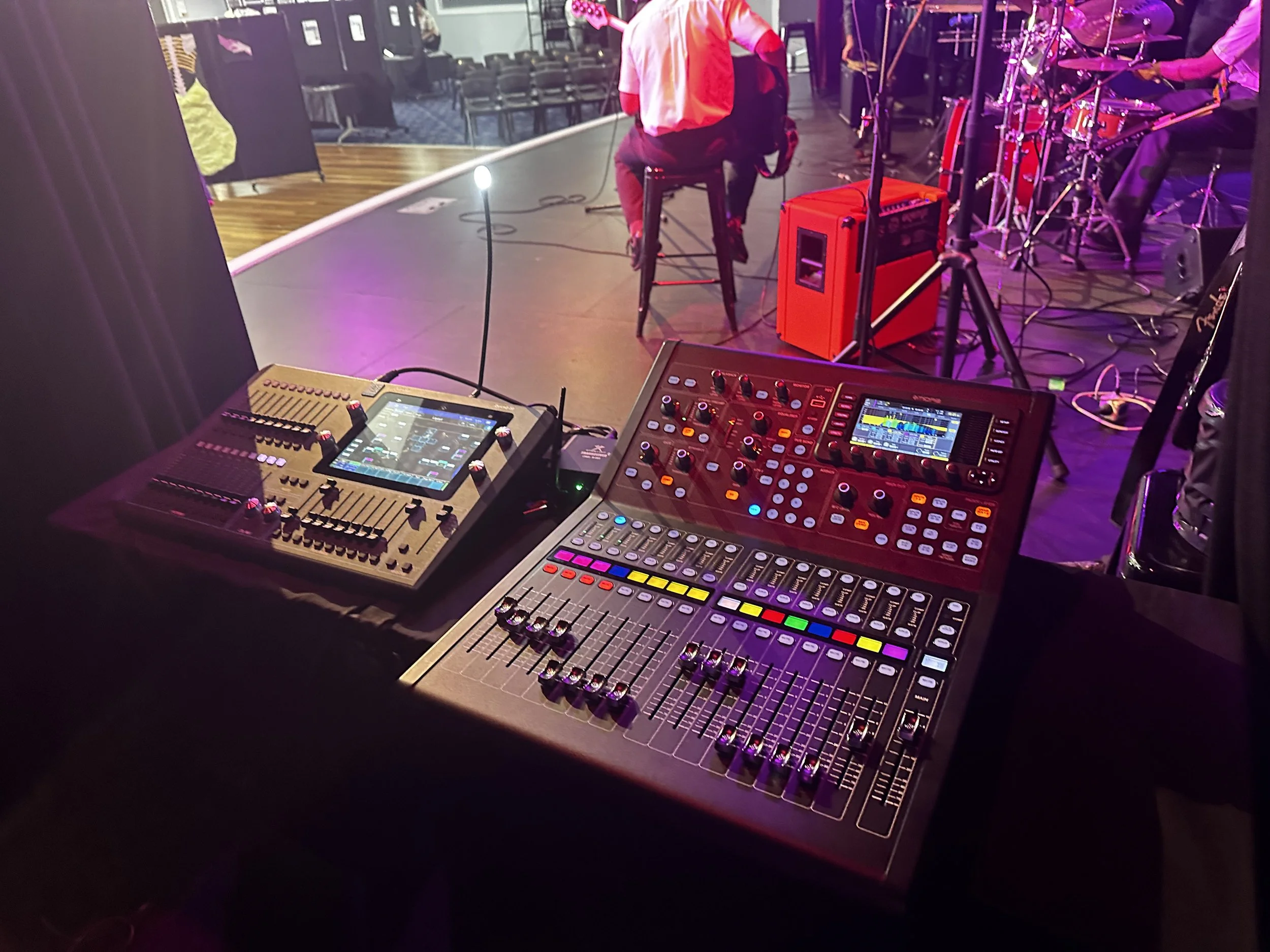 View of a sound mixing console and audio equipment on a table at a live music event. In the background, musicians are playing instruments on stage, with purple and pink lighting.