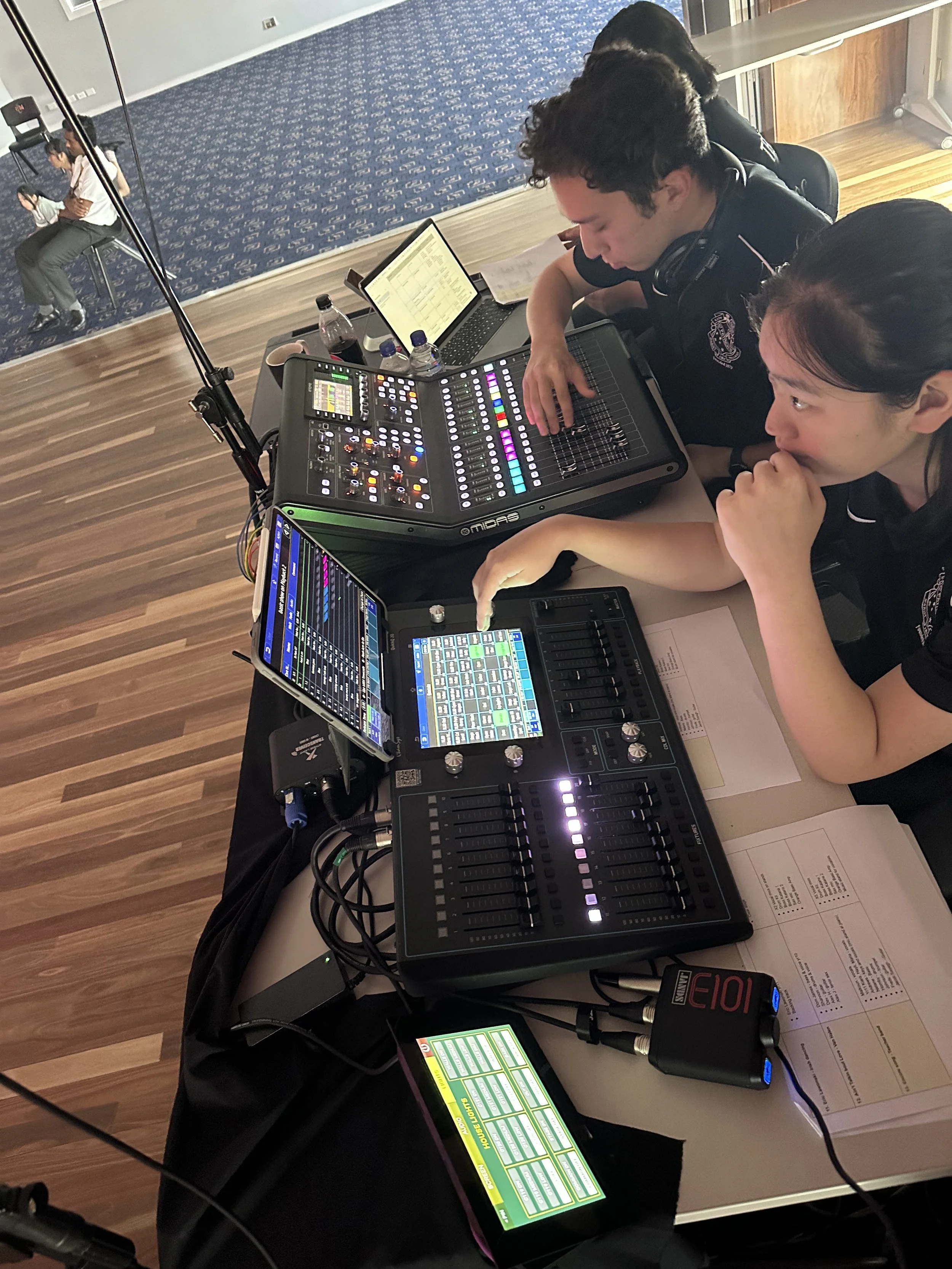 Two women work on sound mixing equipment at a table with digital audio mixers, tablets, and sheets of paper. In the background, a woman plays a cello on a stage with a blue patterned carpet.