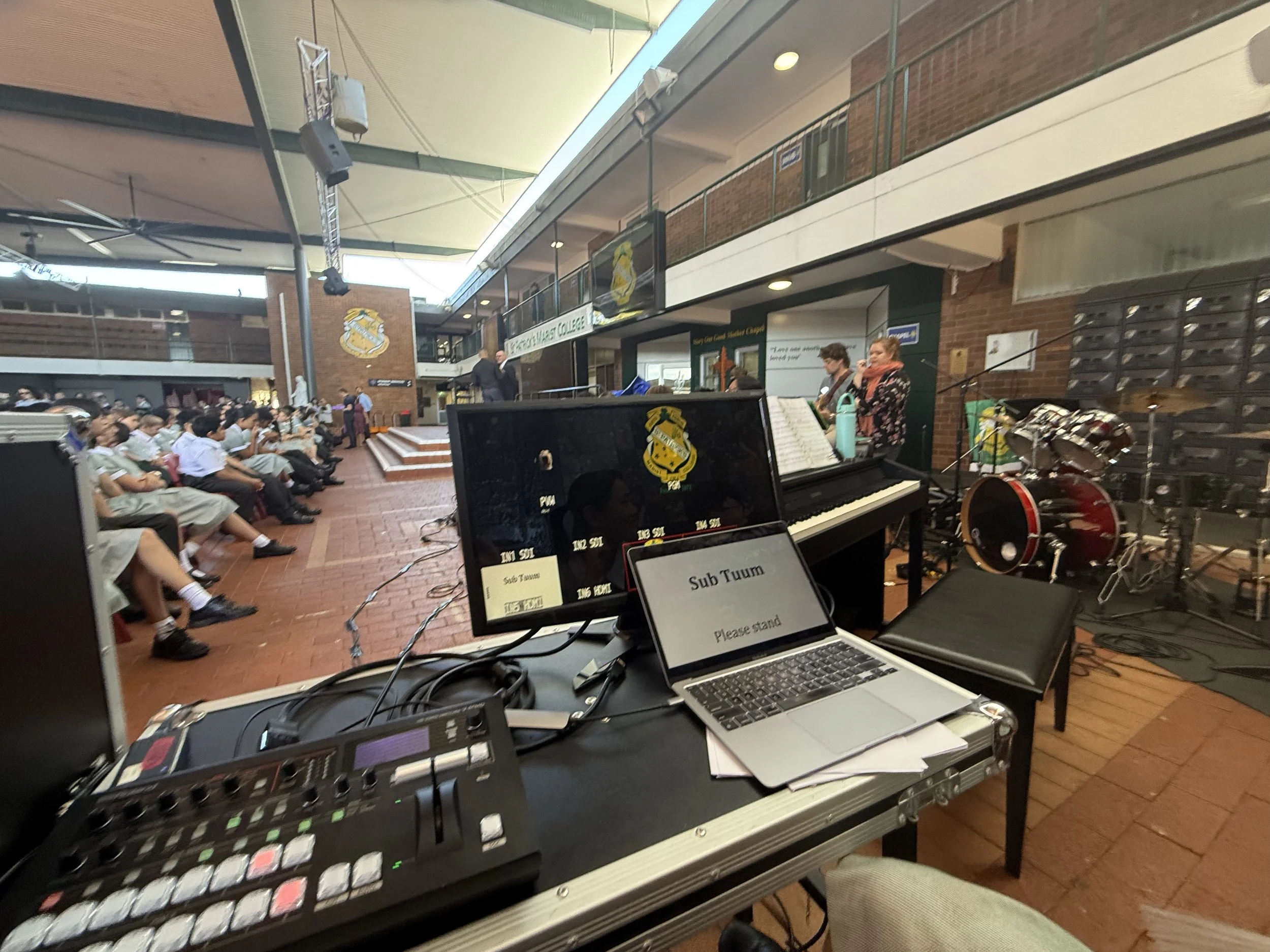 School auditorium with students seated and a band performing on stage. Music equipment, including keyboard, drums, and soundboard, visible in the foreground.