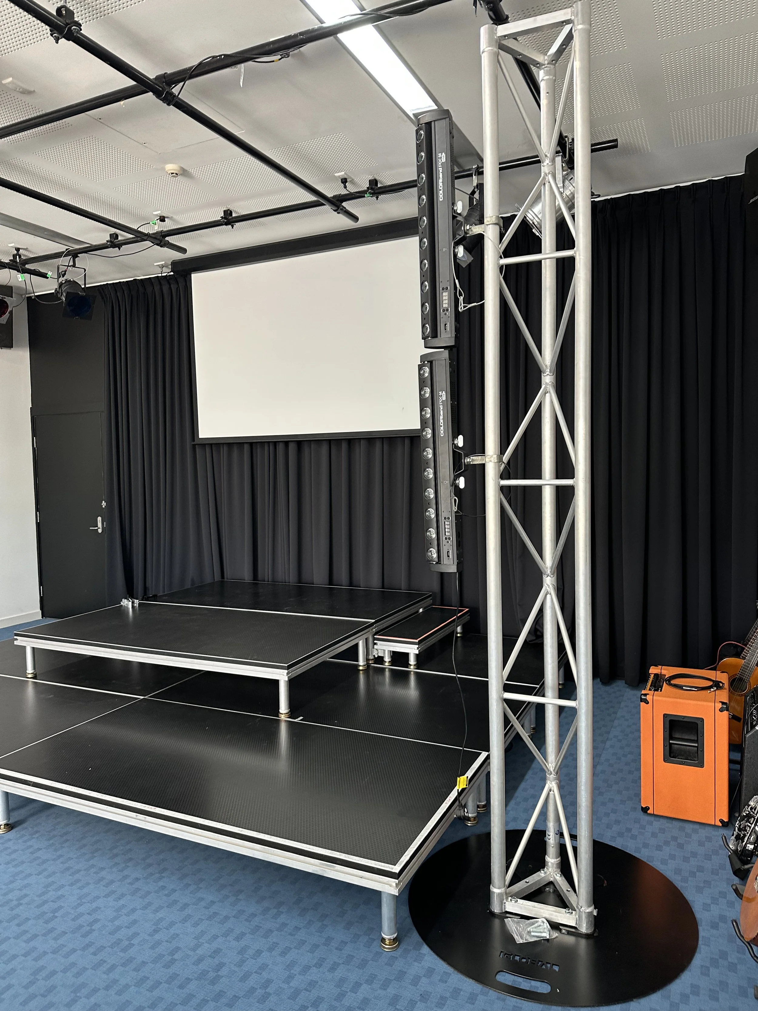 Empty stage with black flooring, black curtains, a large white screen, lighting equipment, and a metal truss structure in a room with blue carpet.