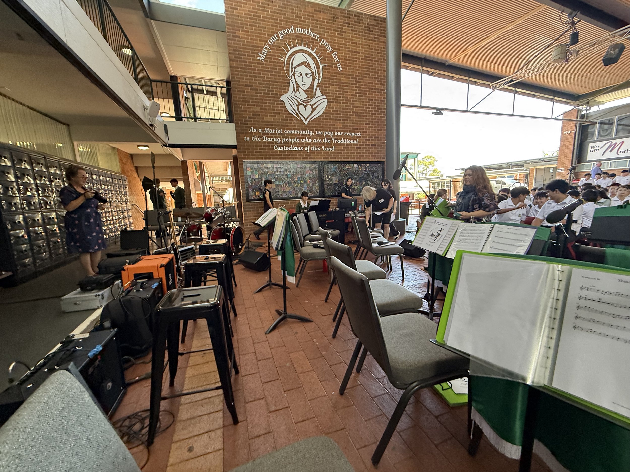An indoor musical performance with musicians and instruments set up, in front of an audience of students in uniforms. A wall features a mural of the Virgin Mary with a quote about respect for traditional custodians. The scene includes chairs, music s