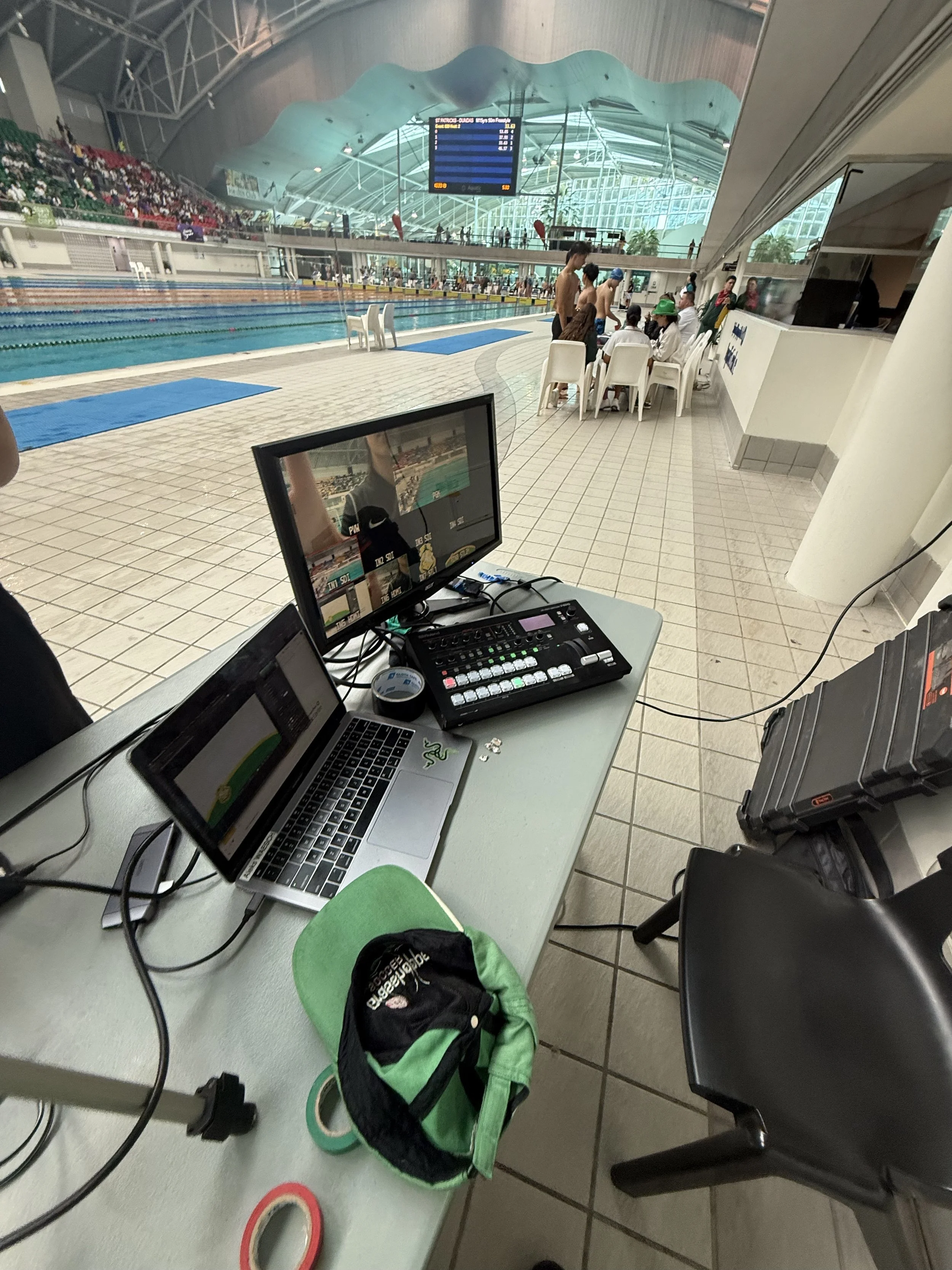 View of an indoor swimming pool area with people sitting and standing near the pool, a digital scoreboard hanging from the ceiling, and a photo booth setup with a laptop, monitor, and various equipment on a table in the foreground.