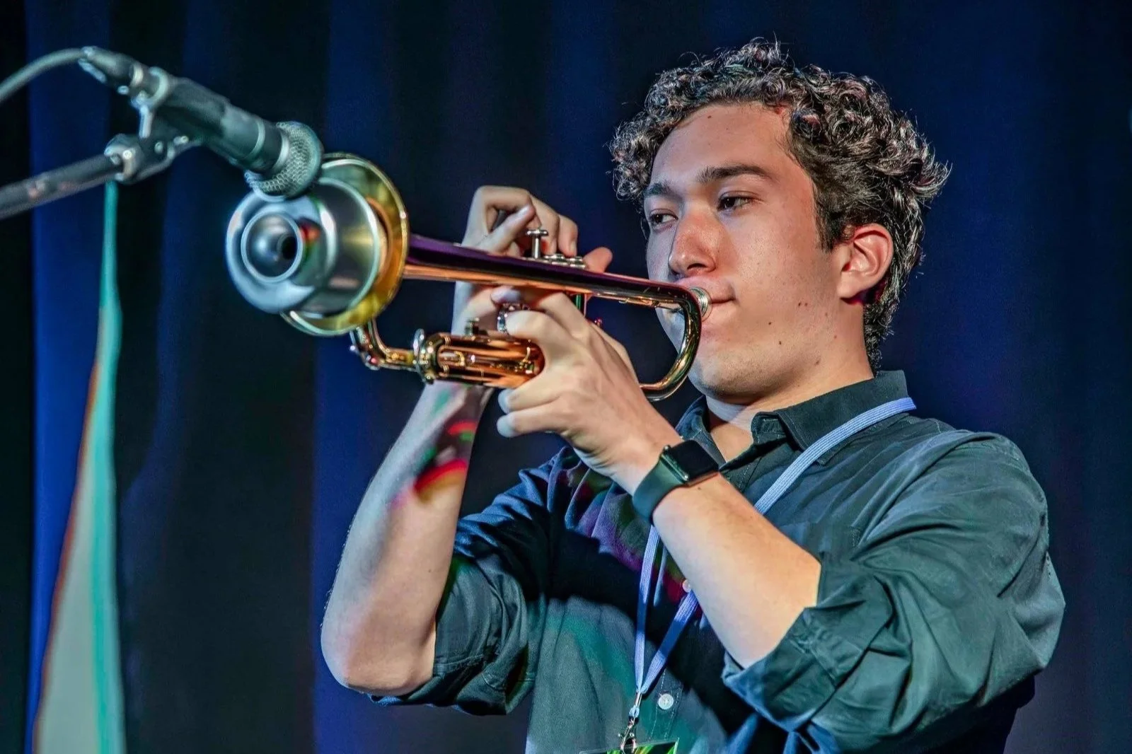 Young man playing trumpet on stage, focused on performance, with dark background and microphone in foreground.