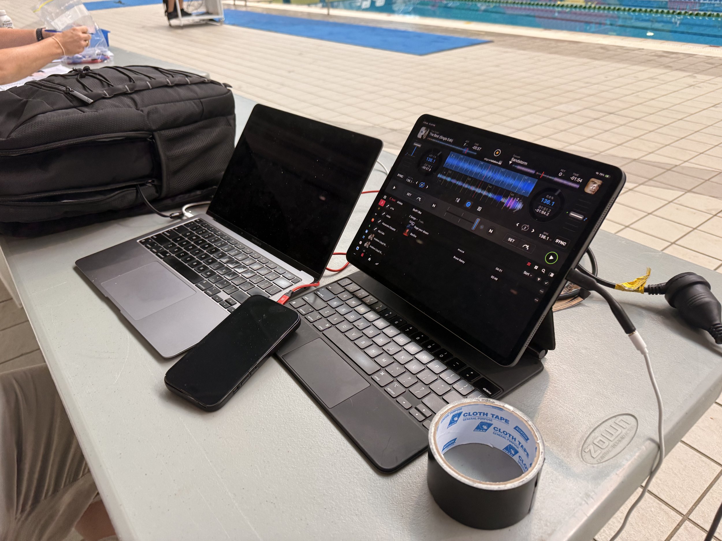 A laptop and a tablet are on a table, with a smartphone, a roll of black tape, and a black backpack nearby. The tablet is displaying a DJ software interface with a music waveform, track controls, and a profile picture. The setting appears to be near 